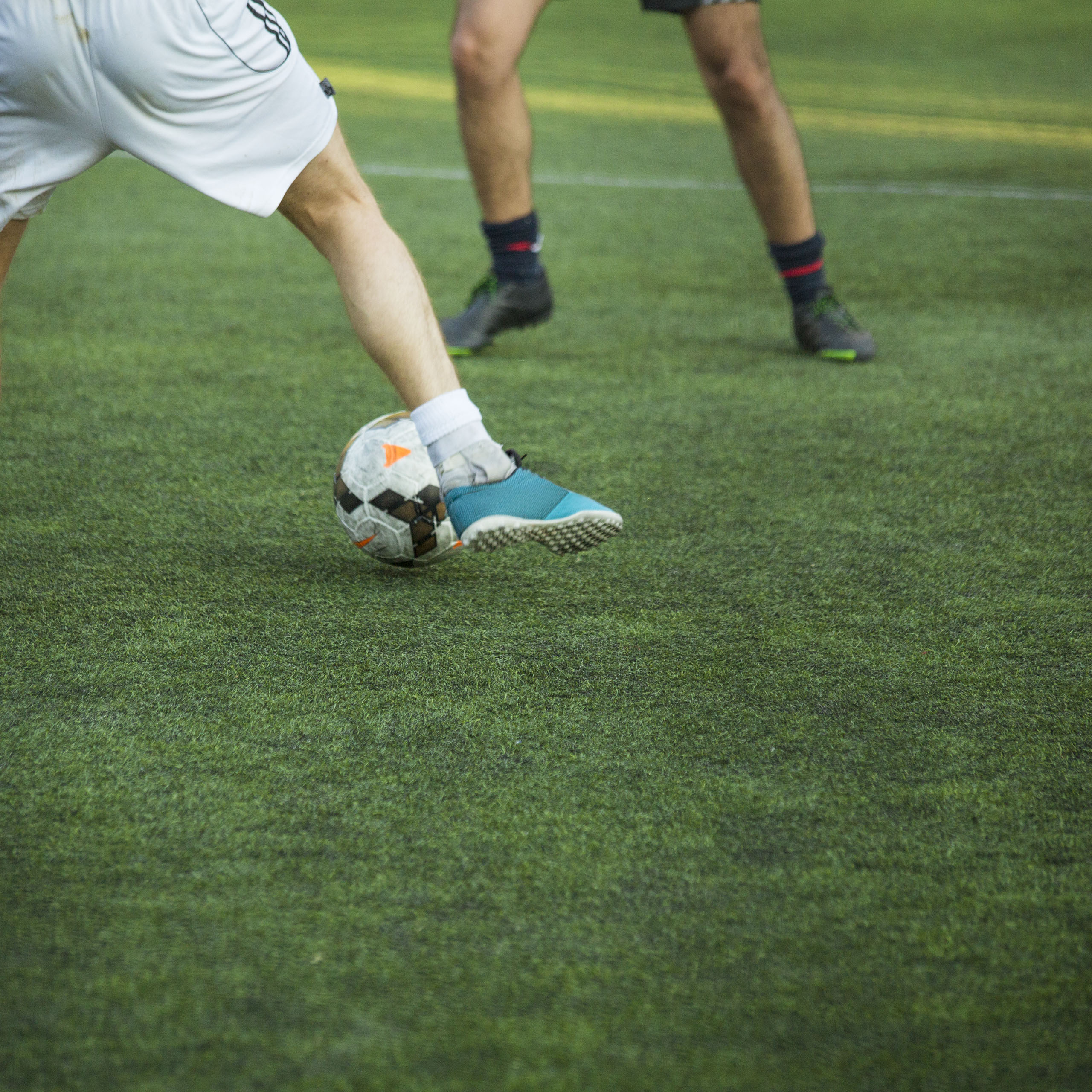 A foot kicks a soccer ball on a field with the legs of another player in the background