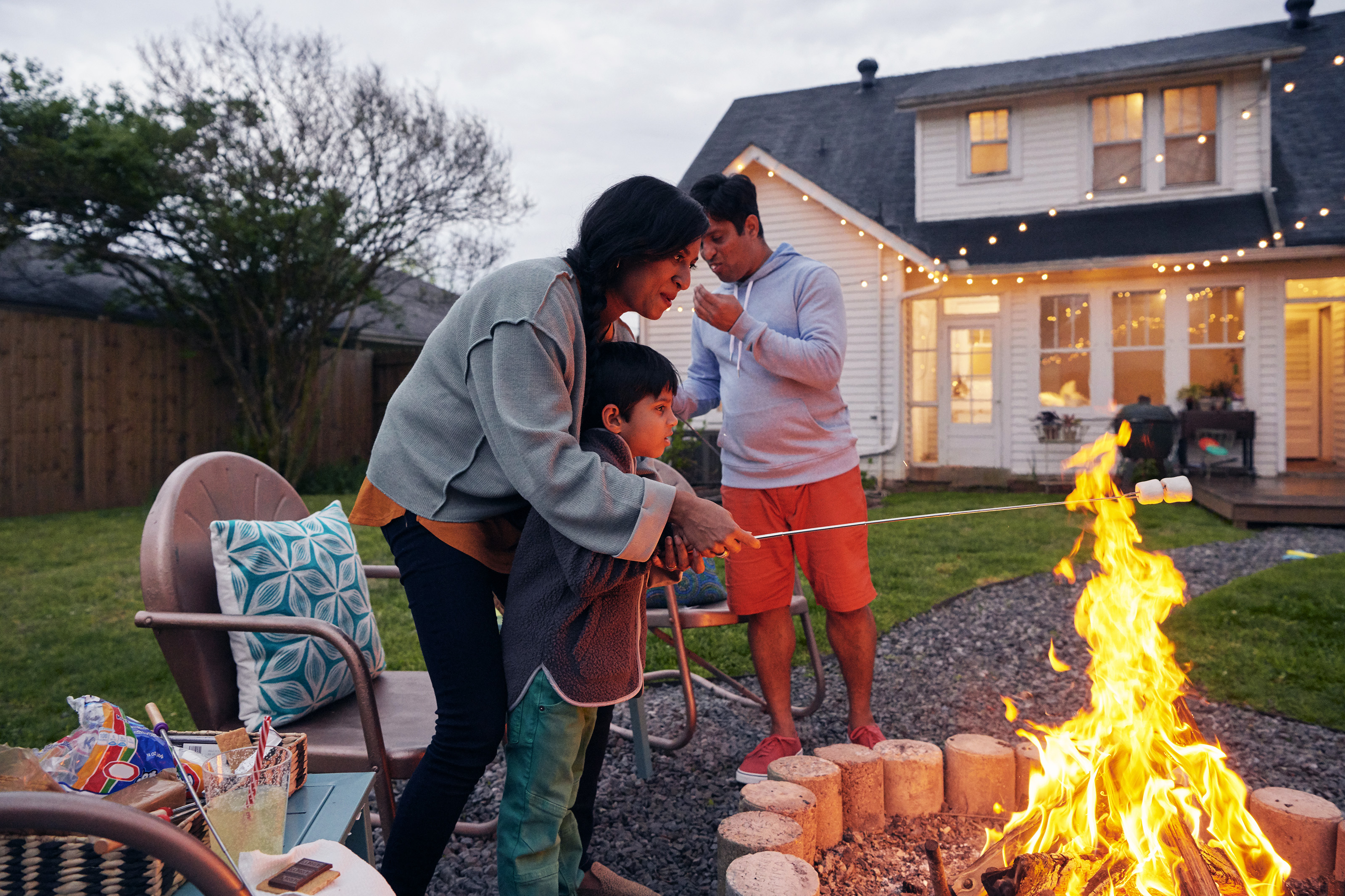Woman cooks s'moors over an open fire with her son, while a man stands in the background in front of a white house.