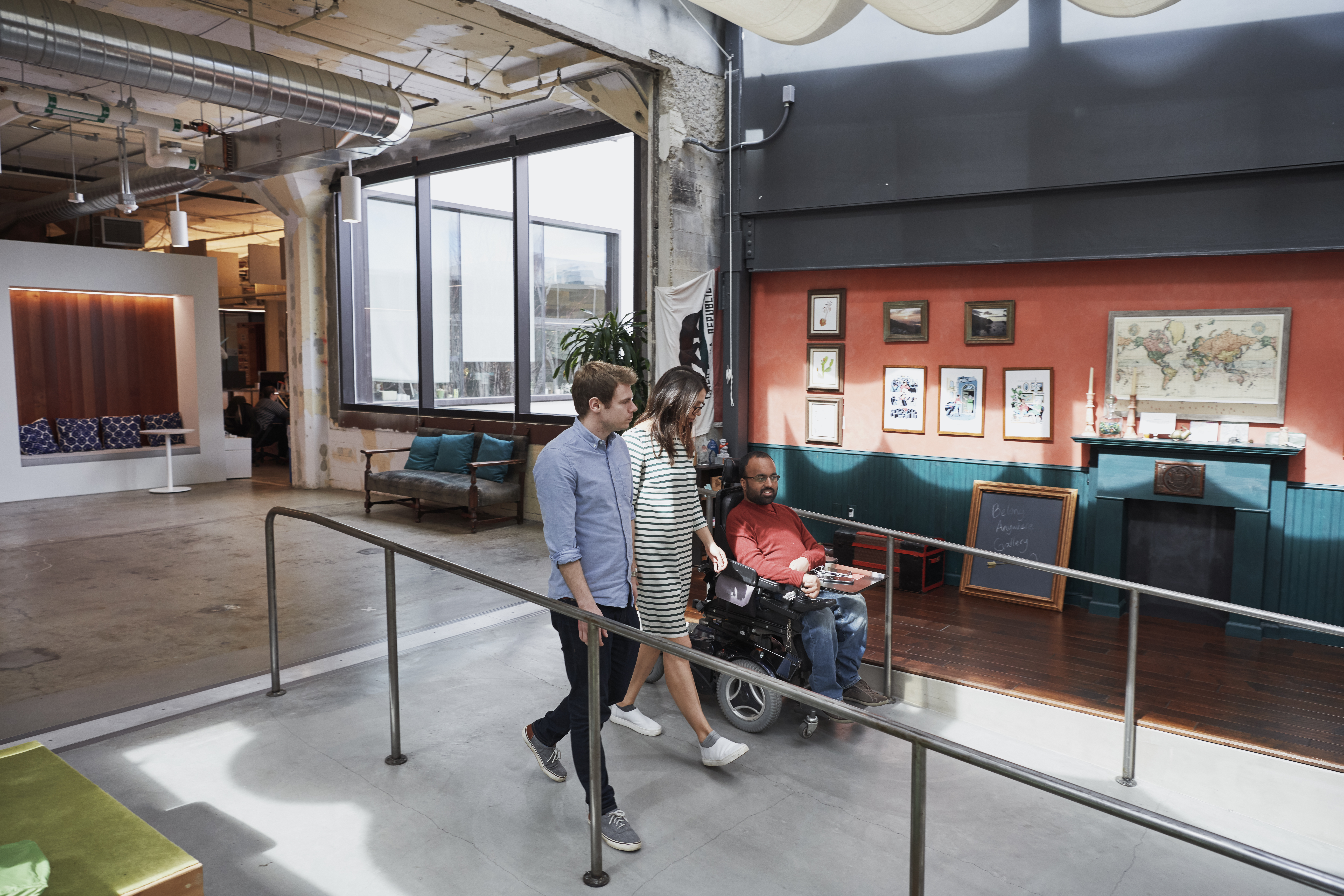 Three people are talking as they walk down a ramp in an office, including a man, a woman and a man using a wheelchair