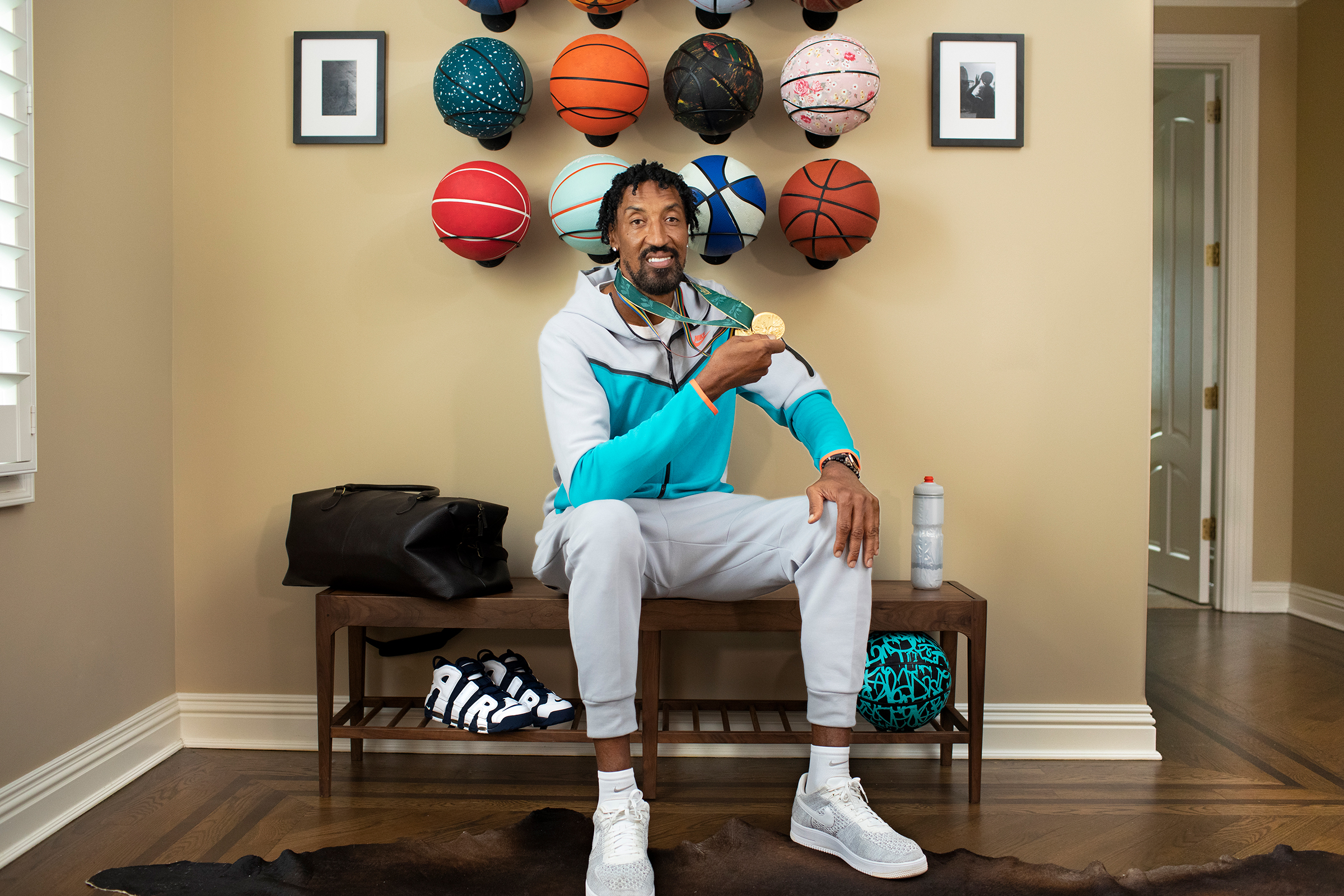 Scottie Pippen sits on a bench in front of basketballs while showing off his 2 Olympic gold medals.
