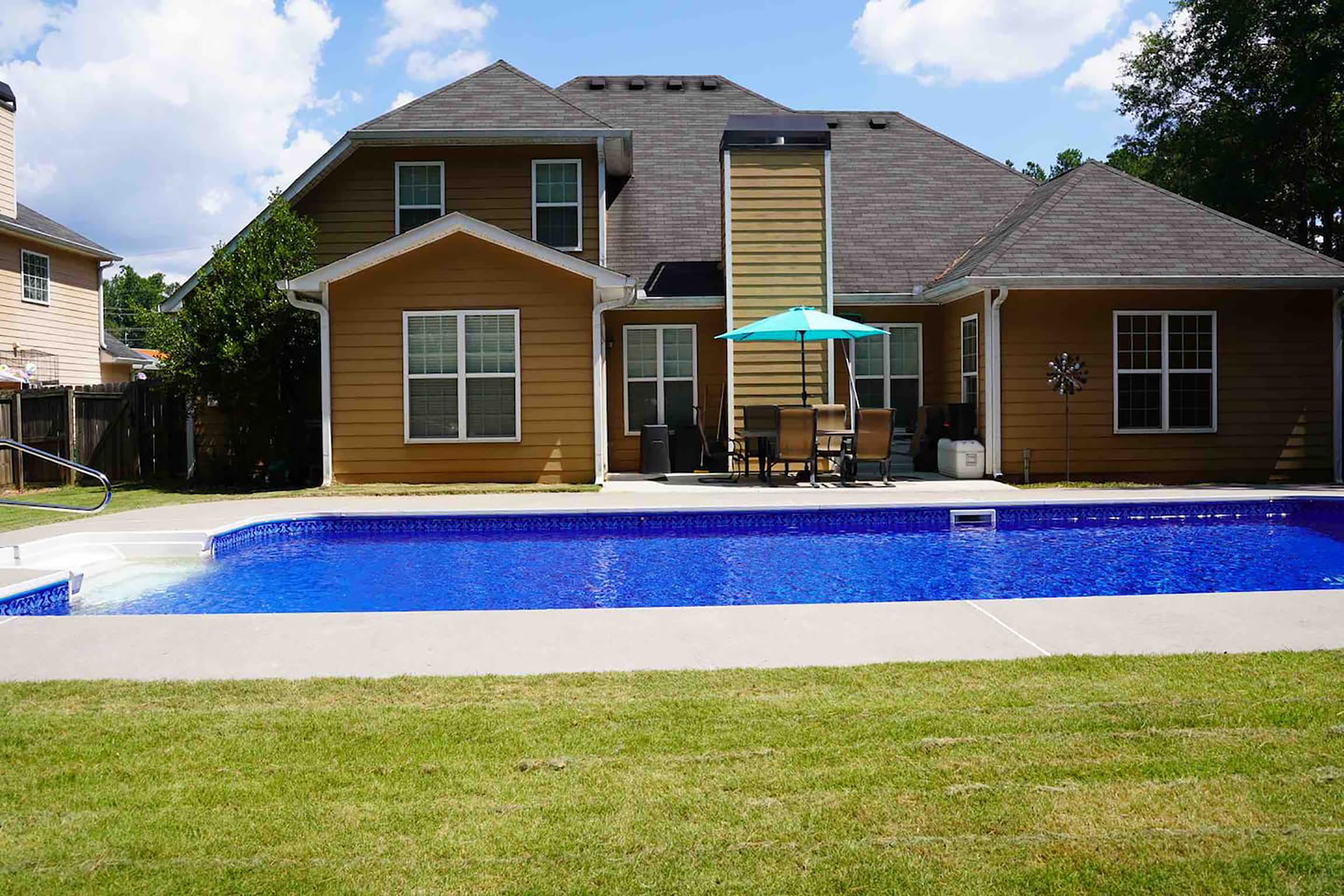 A large, blue swimming pool in front of a tan colored home with outdoor patio furniture 