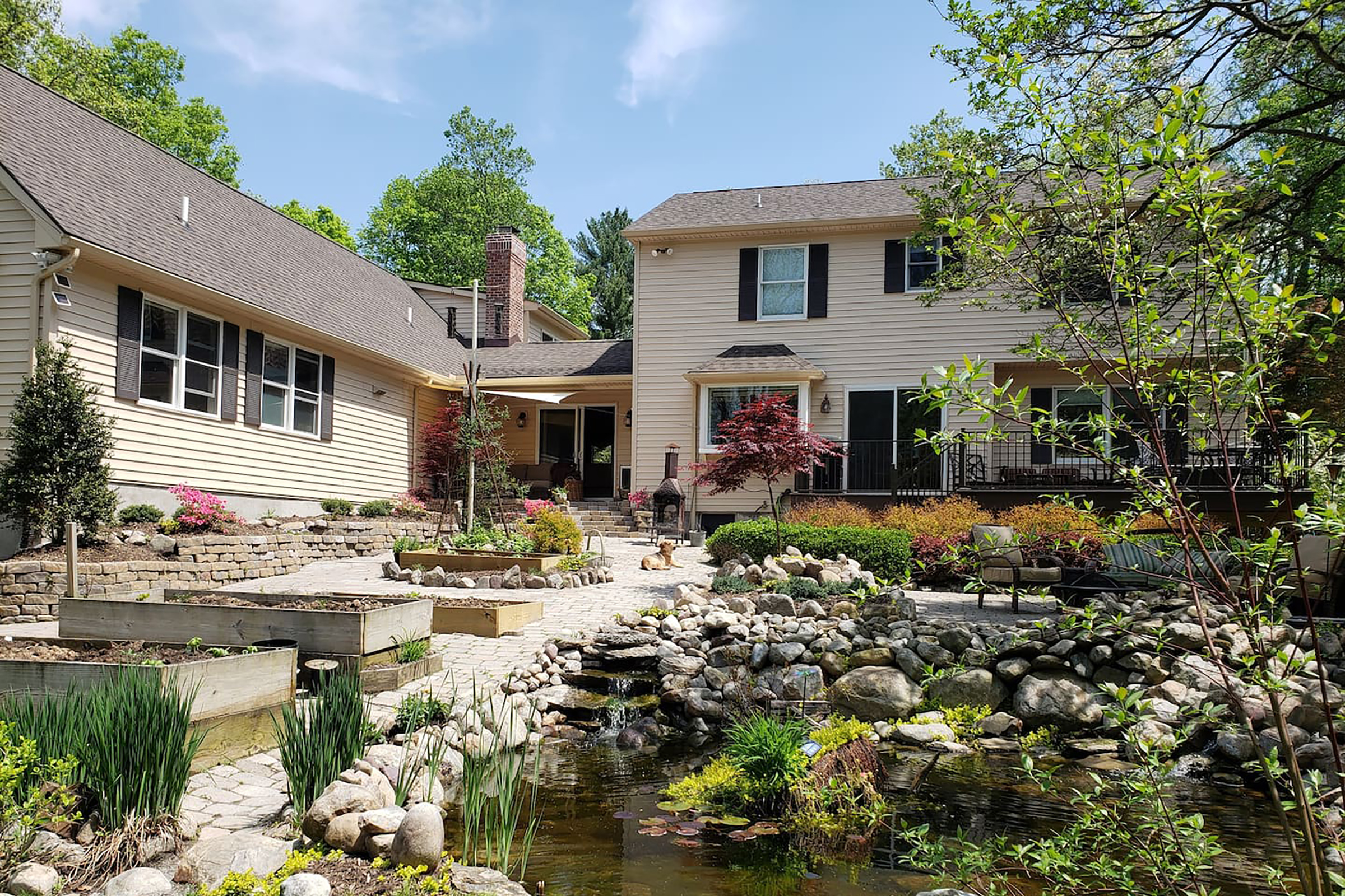 At the forefront is a pond in the backyard of a tan home with a large garden.