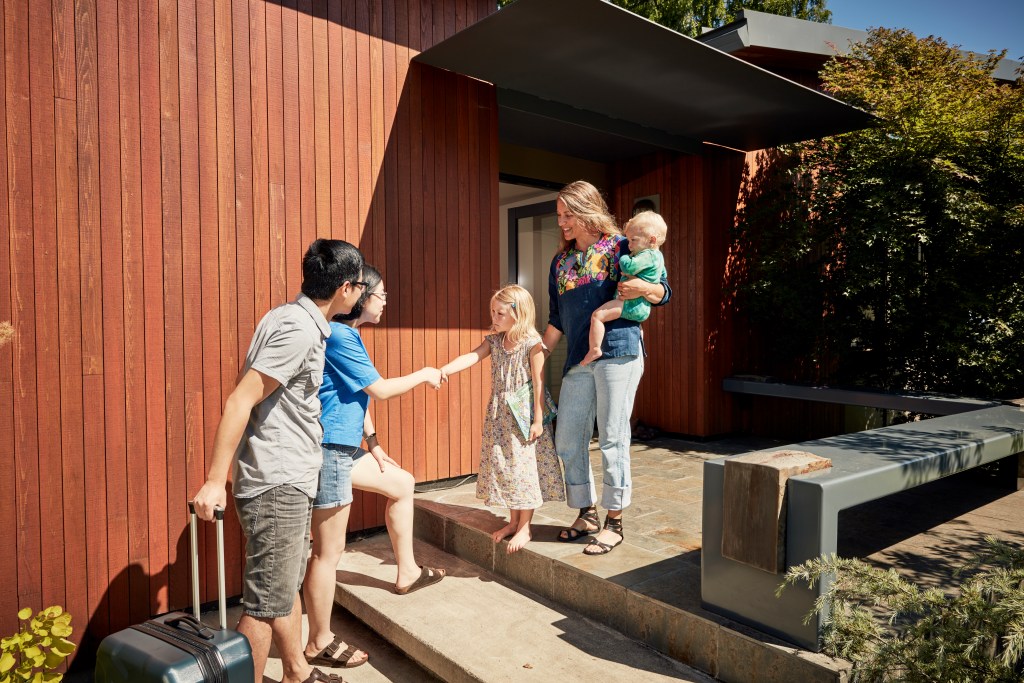 A young Asian couple approach an Airbnb with their luggage, a young mother with her two young children wait to greet them.