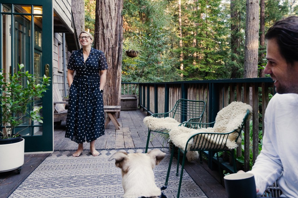 Female guest in blue dress, glasses and pulled back hair stands on a cabin balcony looking at her white dog in the foreground, which is looking back at her. Female's male companion is also seated alongside the dog, wearing a white long sleeved shirt.