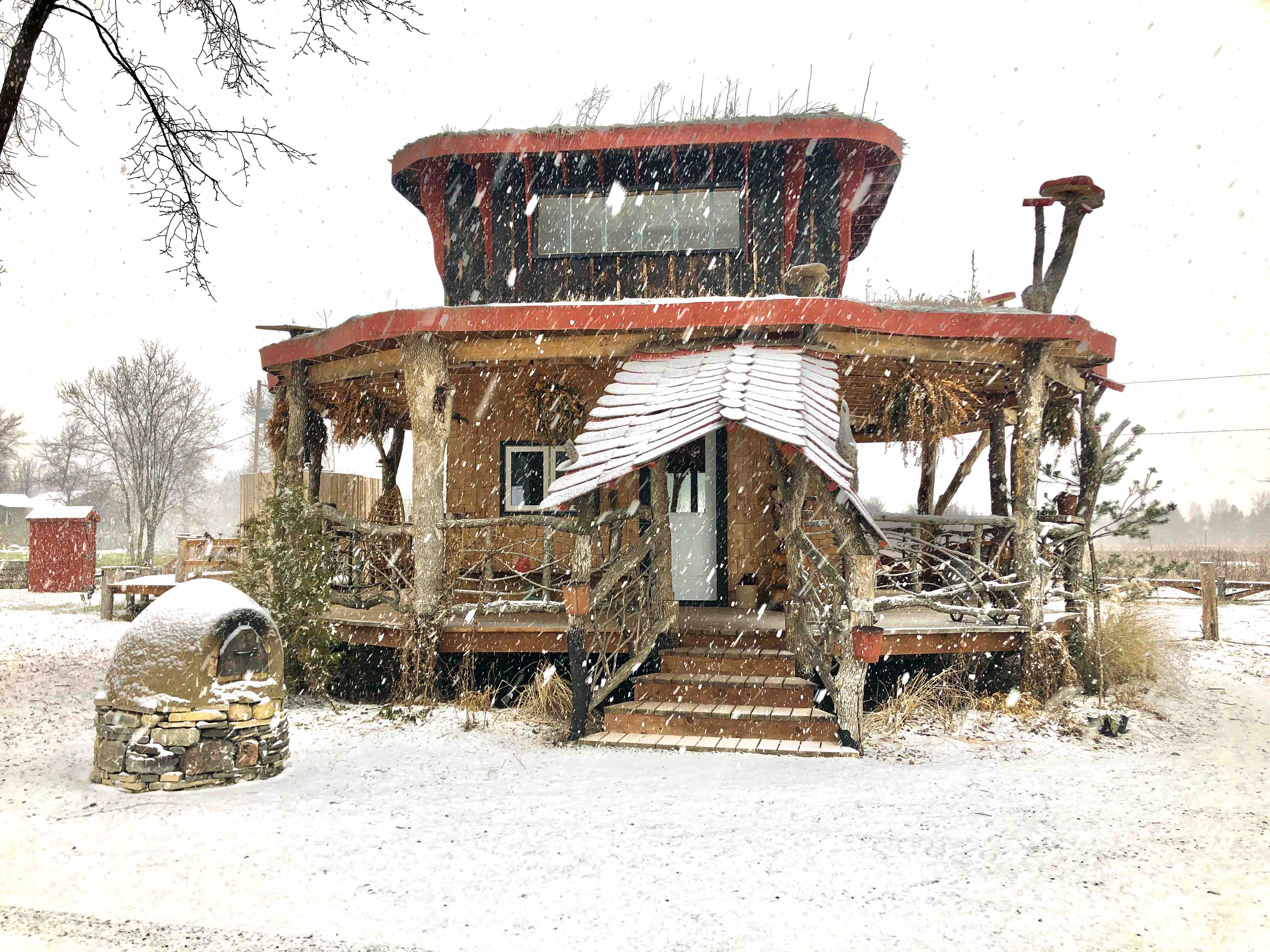 Snow is coming down on a unique wooden house, capped with mushrooms statues.