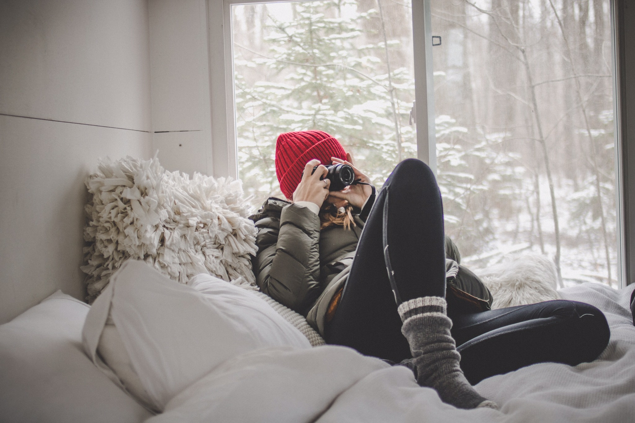 A woman with a red hat and warm comfy clothes lounges on fluffy white bedding with a camera covering her face, preparing to take a photo.
