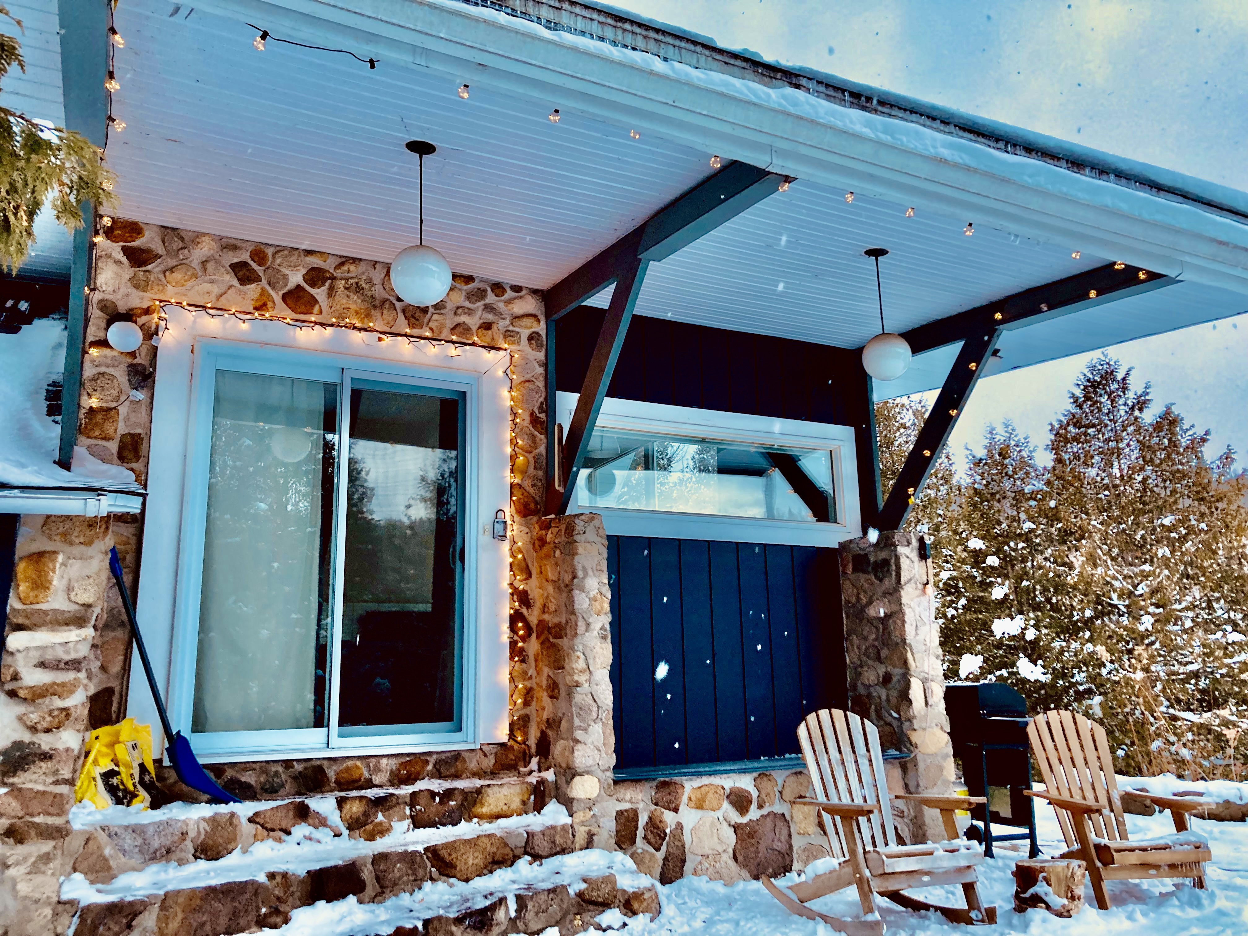 The side door view of a chalet as the snow falls. Two Adirondack chairs sit next to stony steps leading up to a sliding glass door.