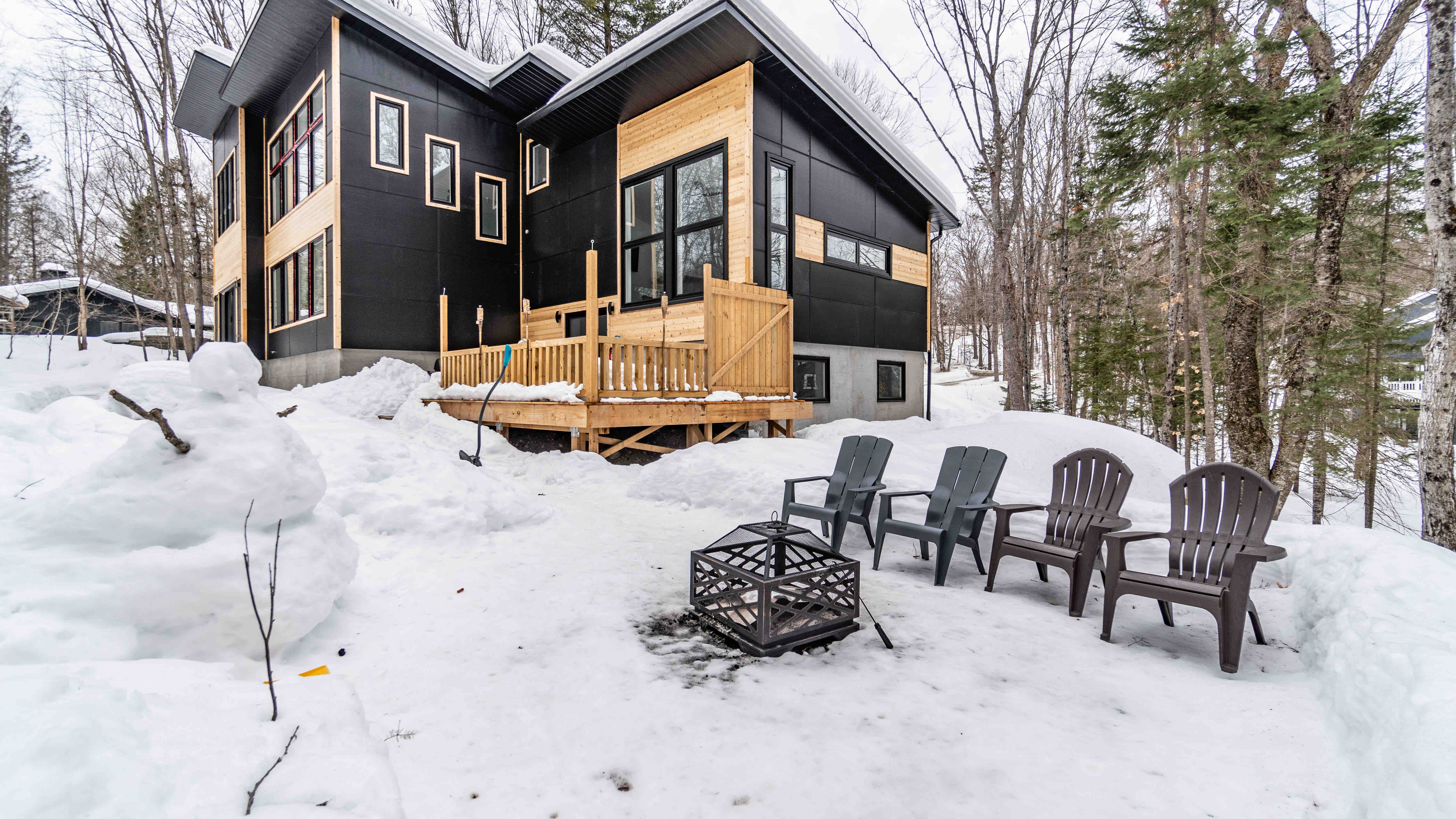 Four outdoor chairs and a firepit are sitting atop snow, with a modern multistory home behind.