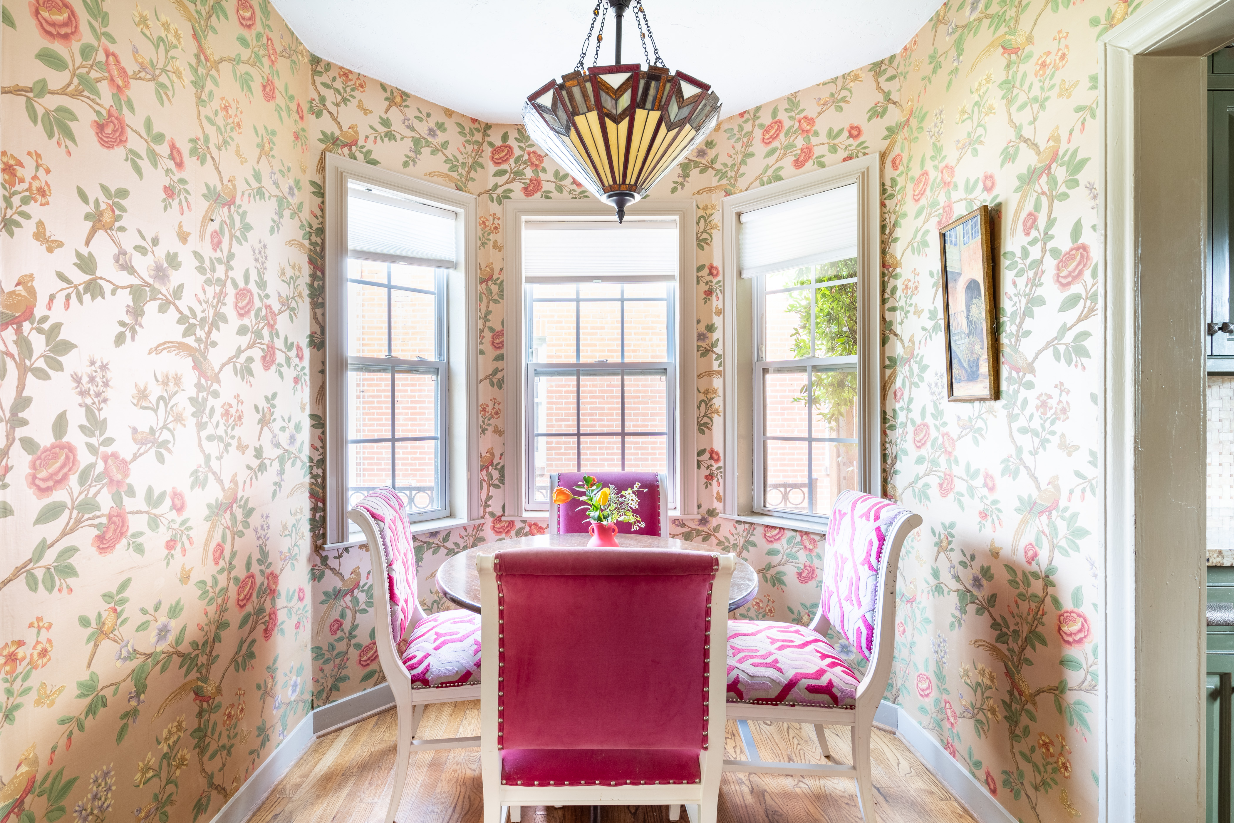 An English country styled breakfast nook, with pink, green and yellow floral wallpaper and a small circular table with pink seating chairs with a bay window in the background.