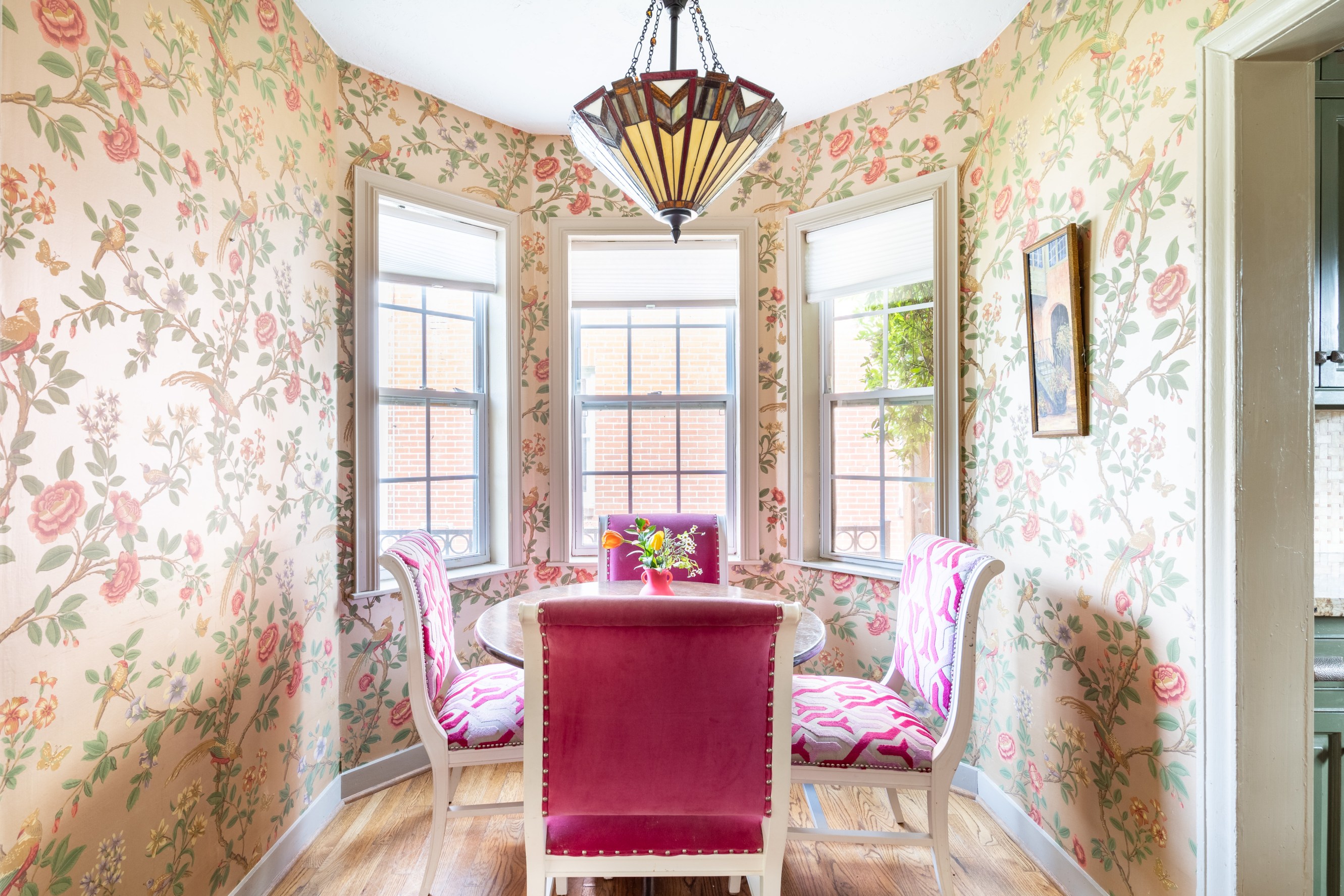 An English country styled breakfast nook, with pink, green and yellow floral wallpaper and a small circular table with pink seating chairs with a bay window in the background.