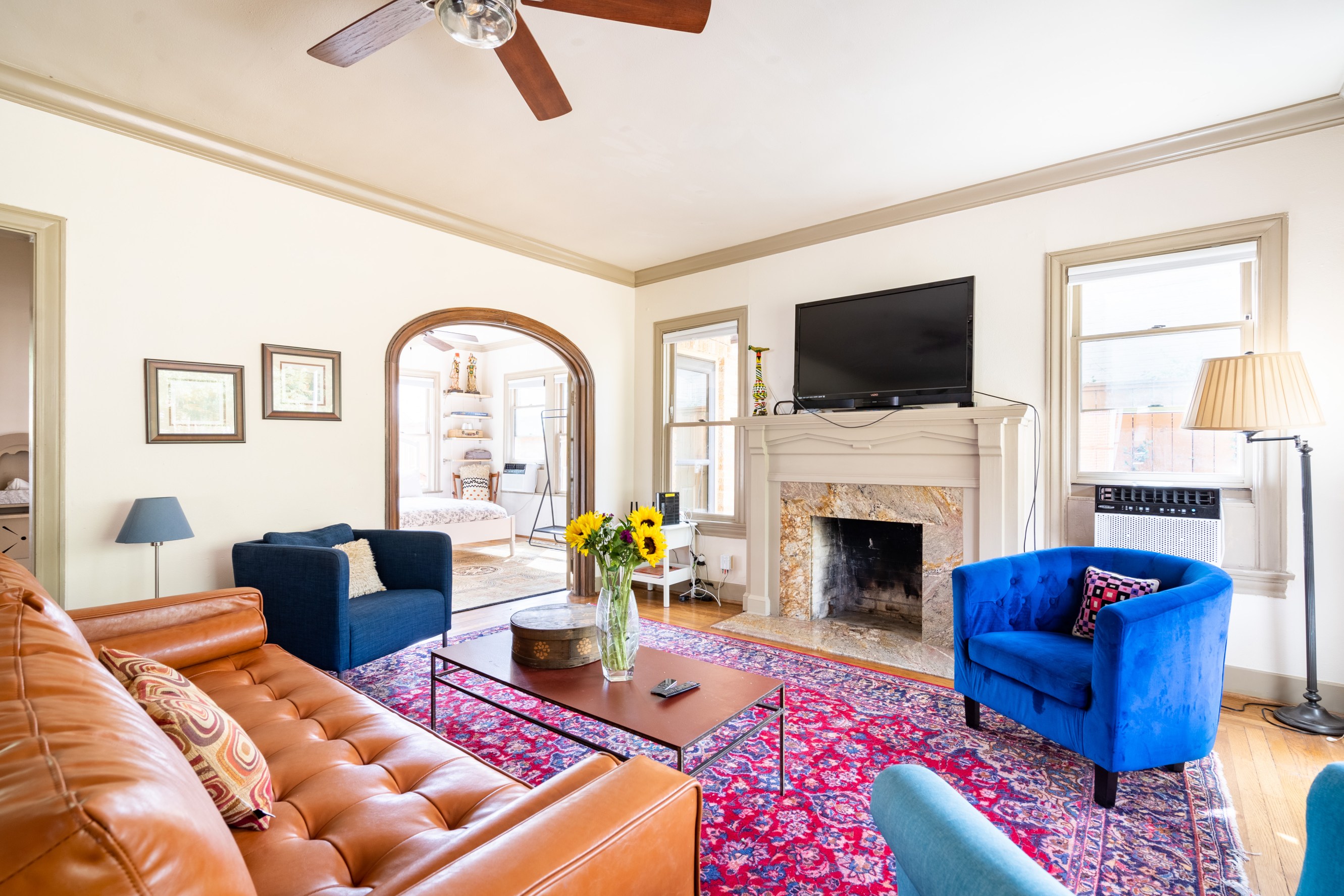 A bright and open living room with an imperial rug, blue accent chairs and a brown leather couch facing a fireplace with a TV above the mantle.
