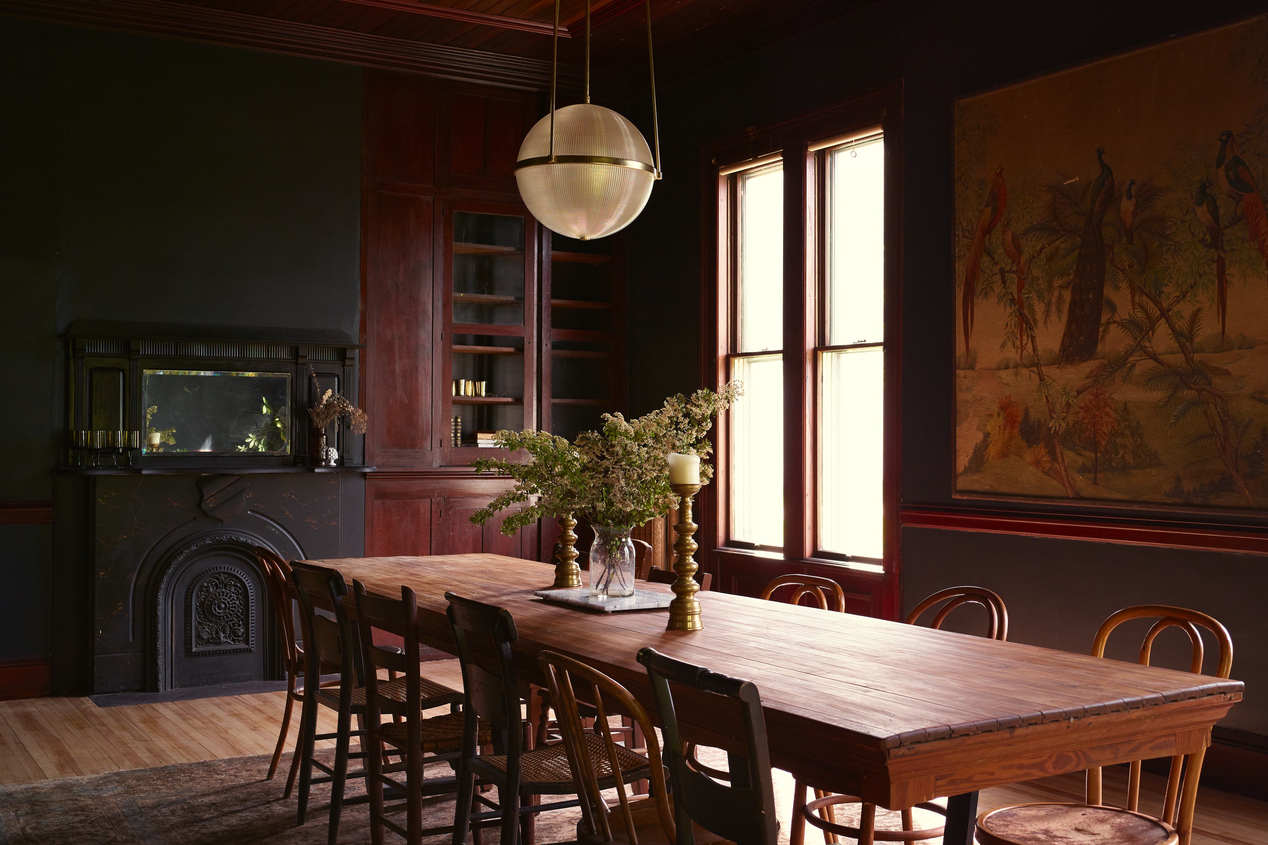 A grand, dark wood dining room with a globe light hanging above the wooden dining table. Flowers and candlesticks serve as the centerpiece with a black painted fireplace in the background and an antique tapestry on the wall.
