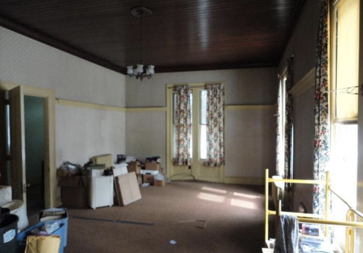 Old empty living room space, dimly lit due to the windows being covered by patterned curtains. Boxes and clutter seen in the back corner.