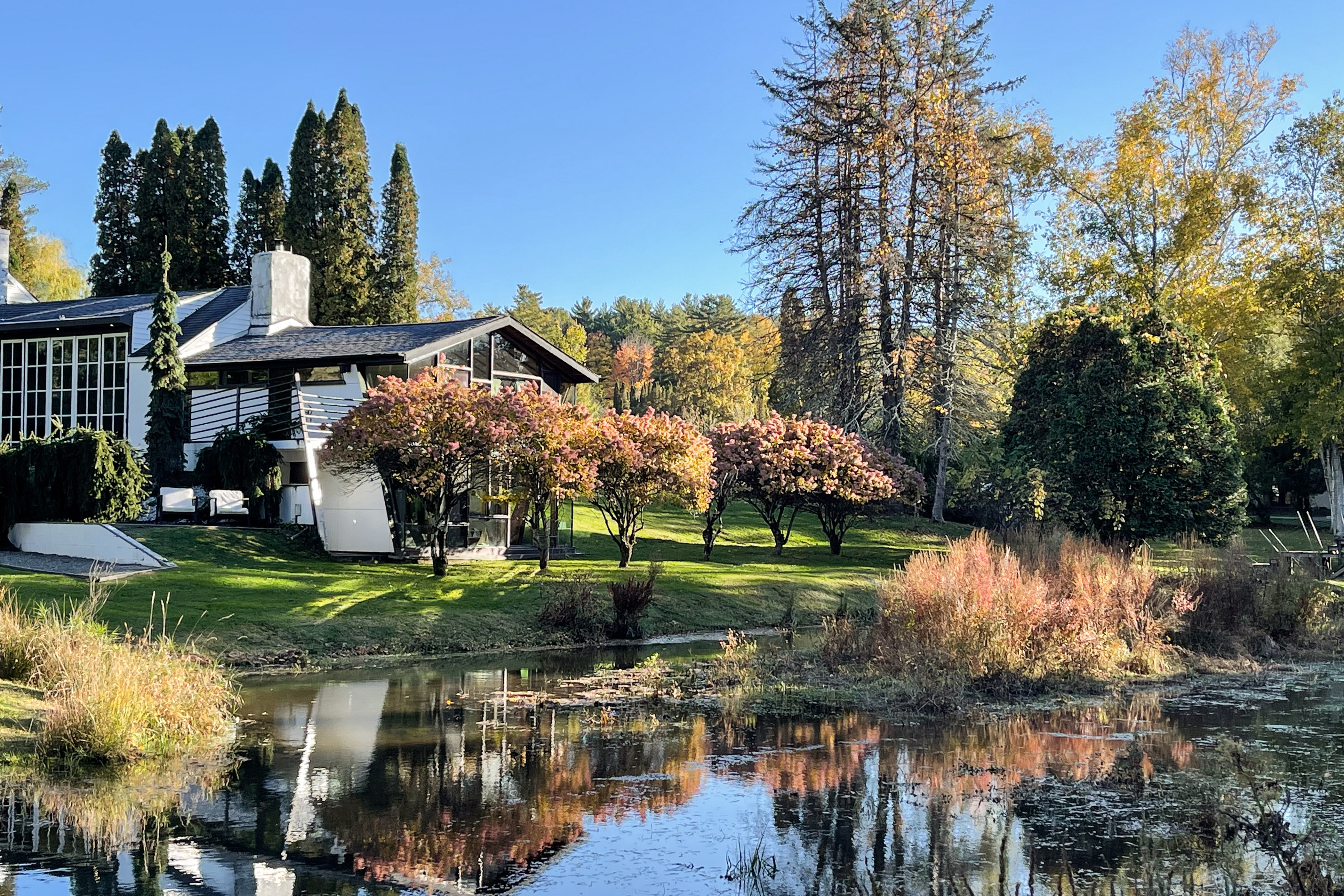 The Pond House property in the background with a view of the yard and pond surrounded by fall foliage.
