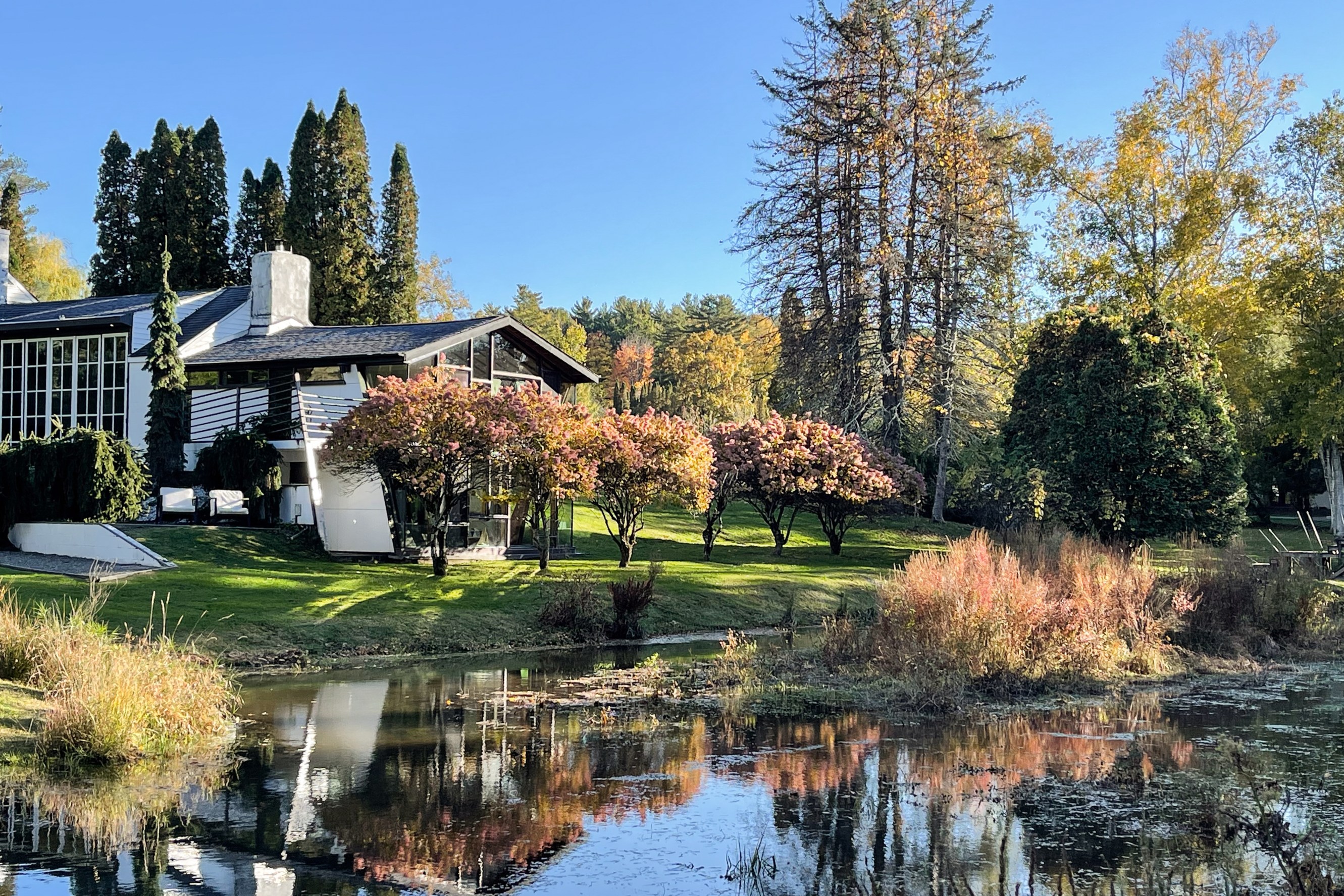 The Pond House property in the background with a view of the yard and pond surrounded by fall foliage.