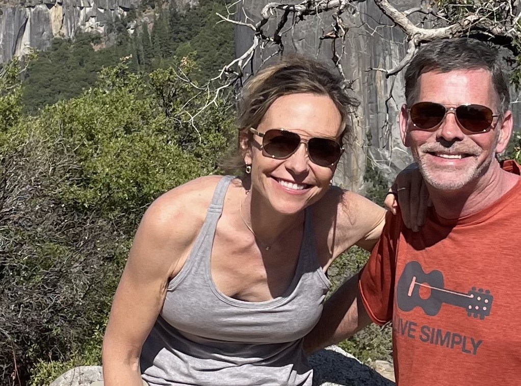 Man and woman smiling for a photo outside with a scenic nature and mountain background. 
