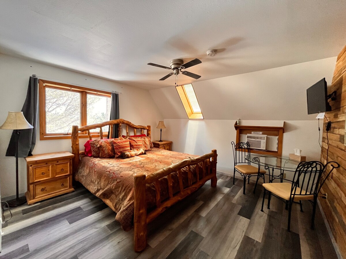 A bedroom with a double bed, ceiling fan, tv and two sitting chairs with table.