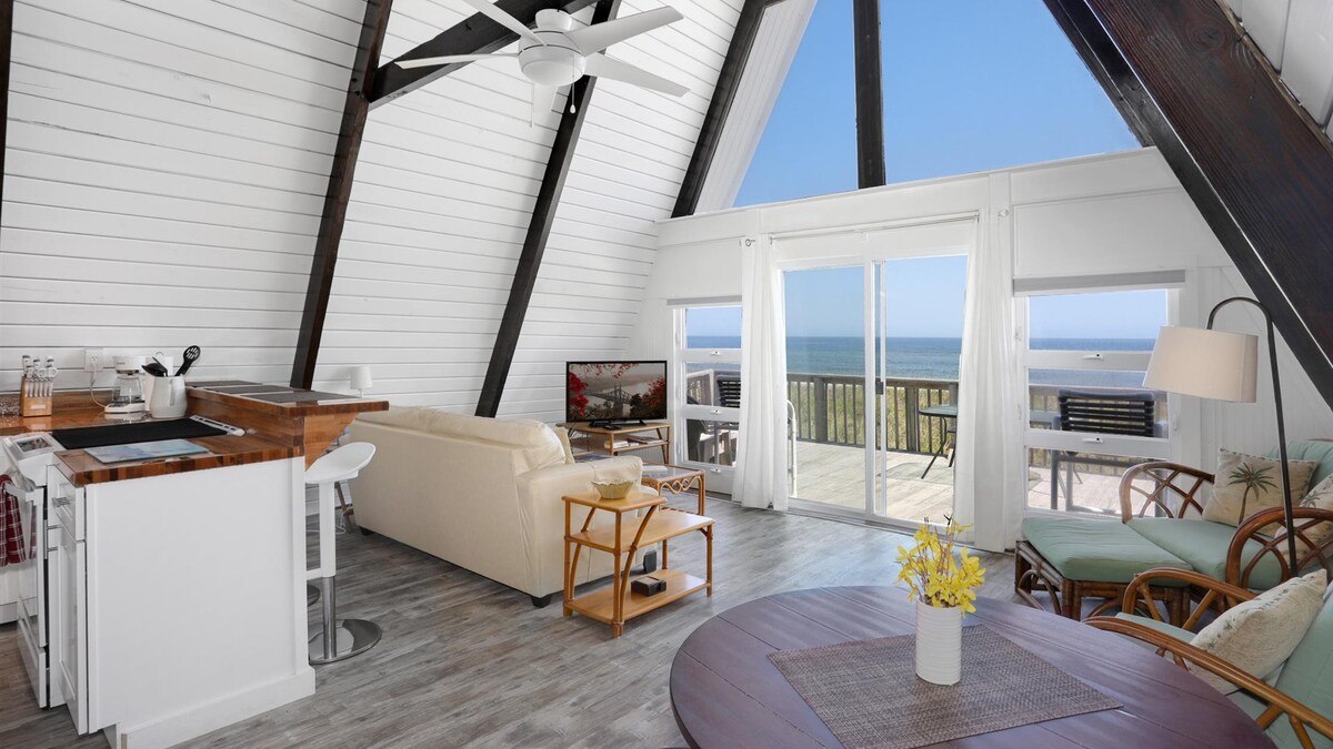 An a-frame cabin sitting room, with a couch - tv - lamp - looking onto the decking and out to the view of the ocean. 