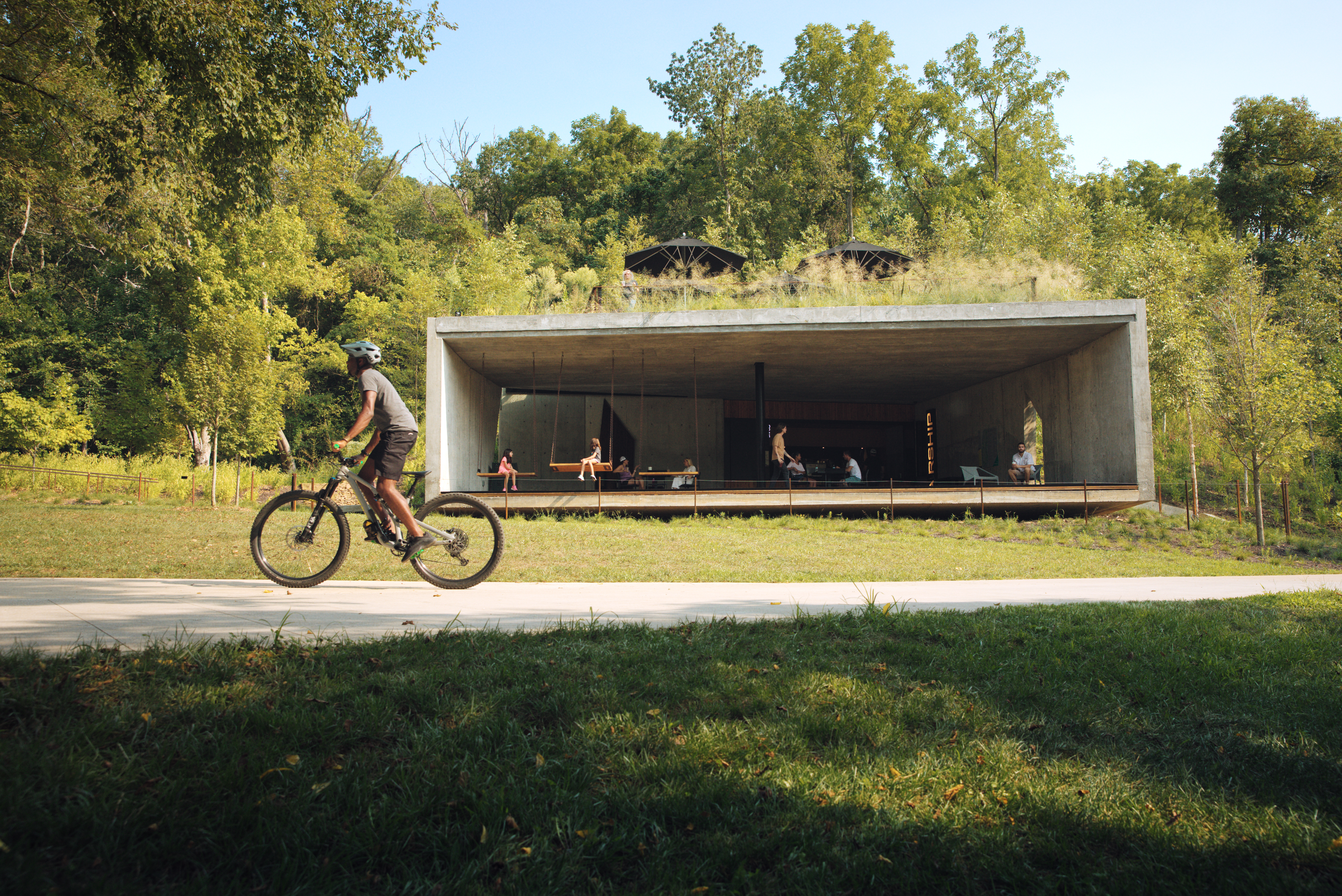 Man rides bike in front of grass field with a concrete container ship in the background
