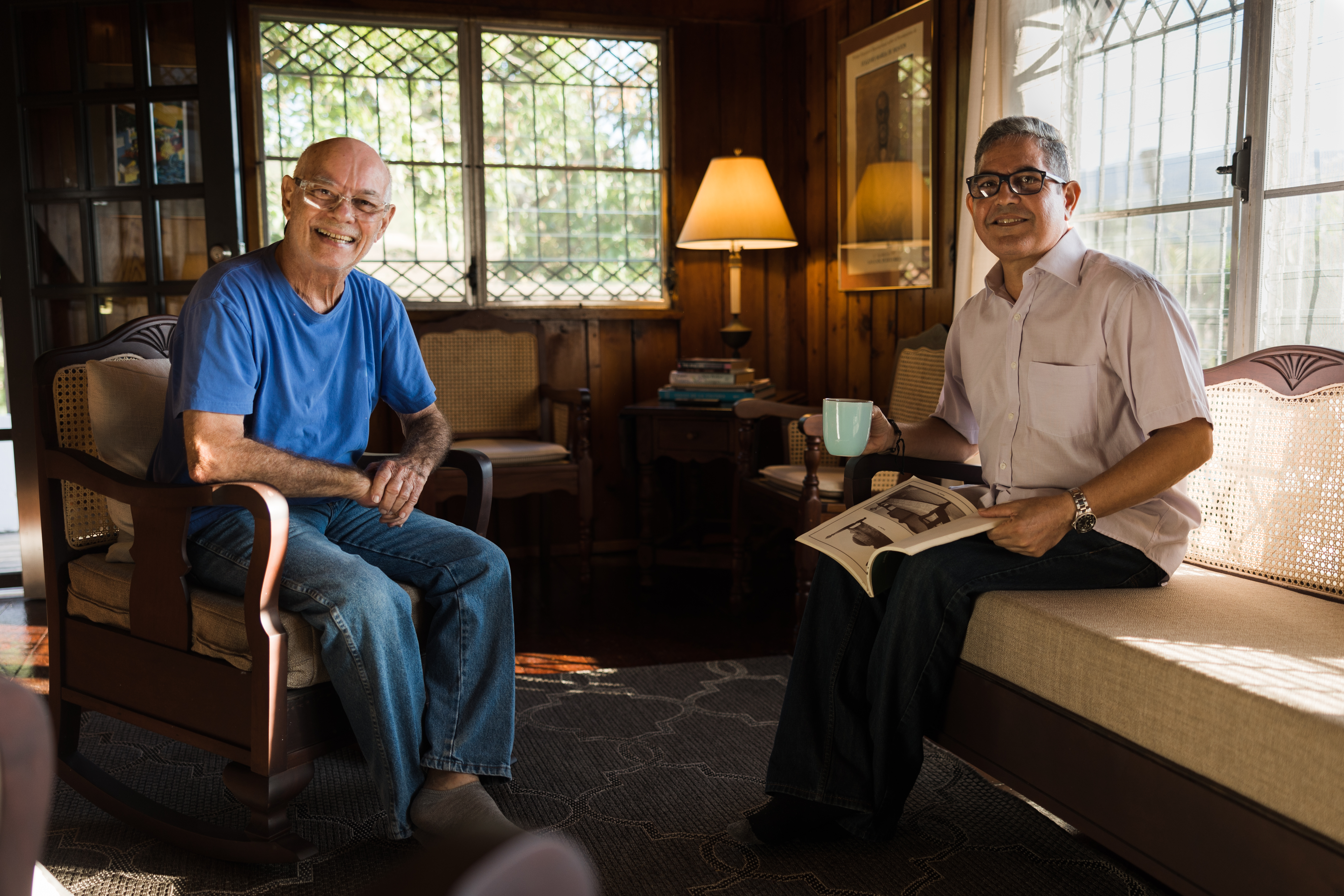 Dos anfitriones de la plataforma están sentados en la sala de la casa, con dos ventanas al fondo.