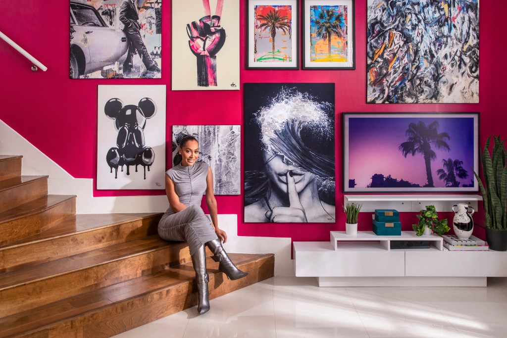 Woman in a grey dress and silver boots smiling and sitting at the bottom of a wooden staircase with a gallery wall of art behind her on a bright pink wall.