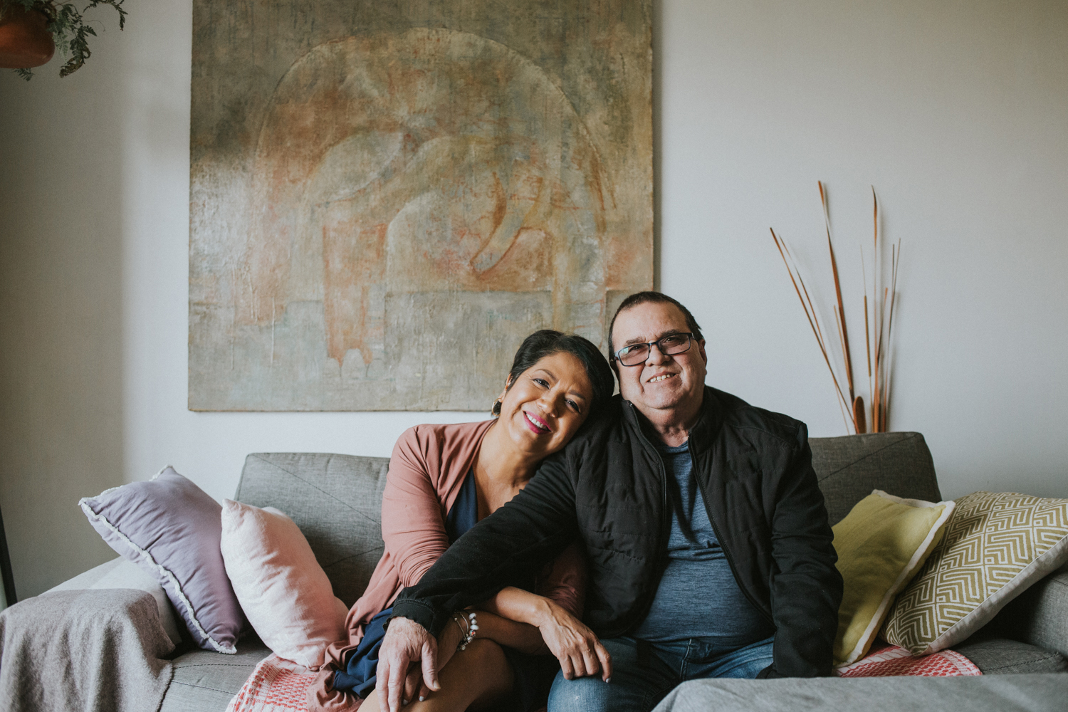 Una pareja sentada en un sofa de sala, mirando de frente a la camara. La mujer recuesta su cabeza en el hombro del ombre