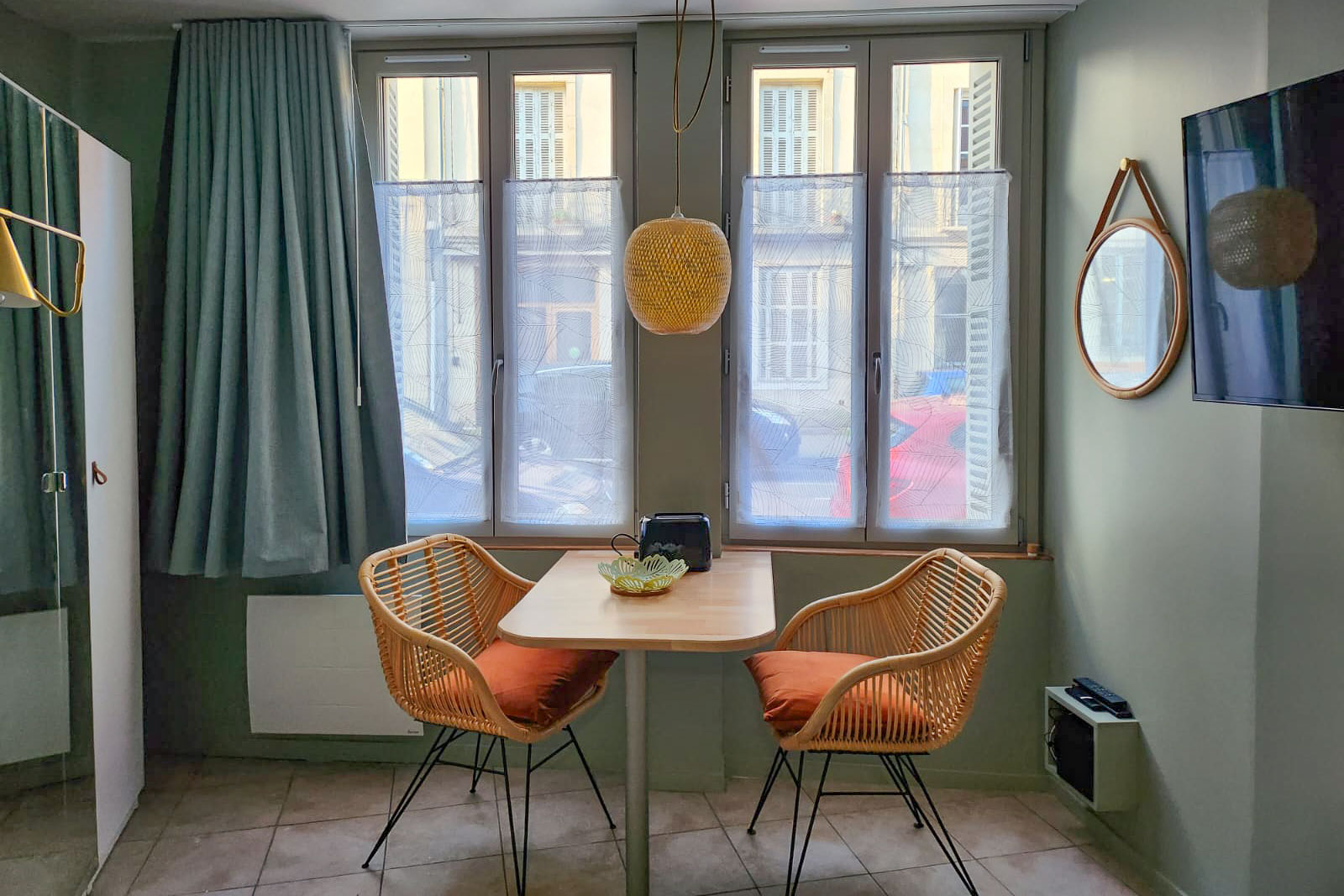 A small table and two wicker chairs in front of a first-floor window, backed by day lit windows and sage green walls. 