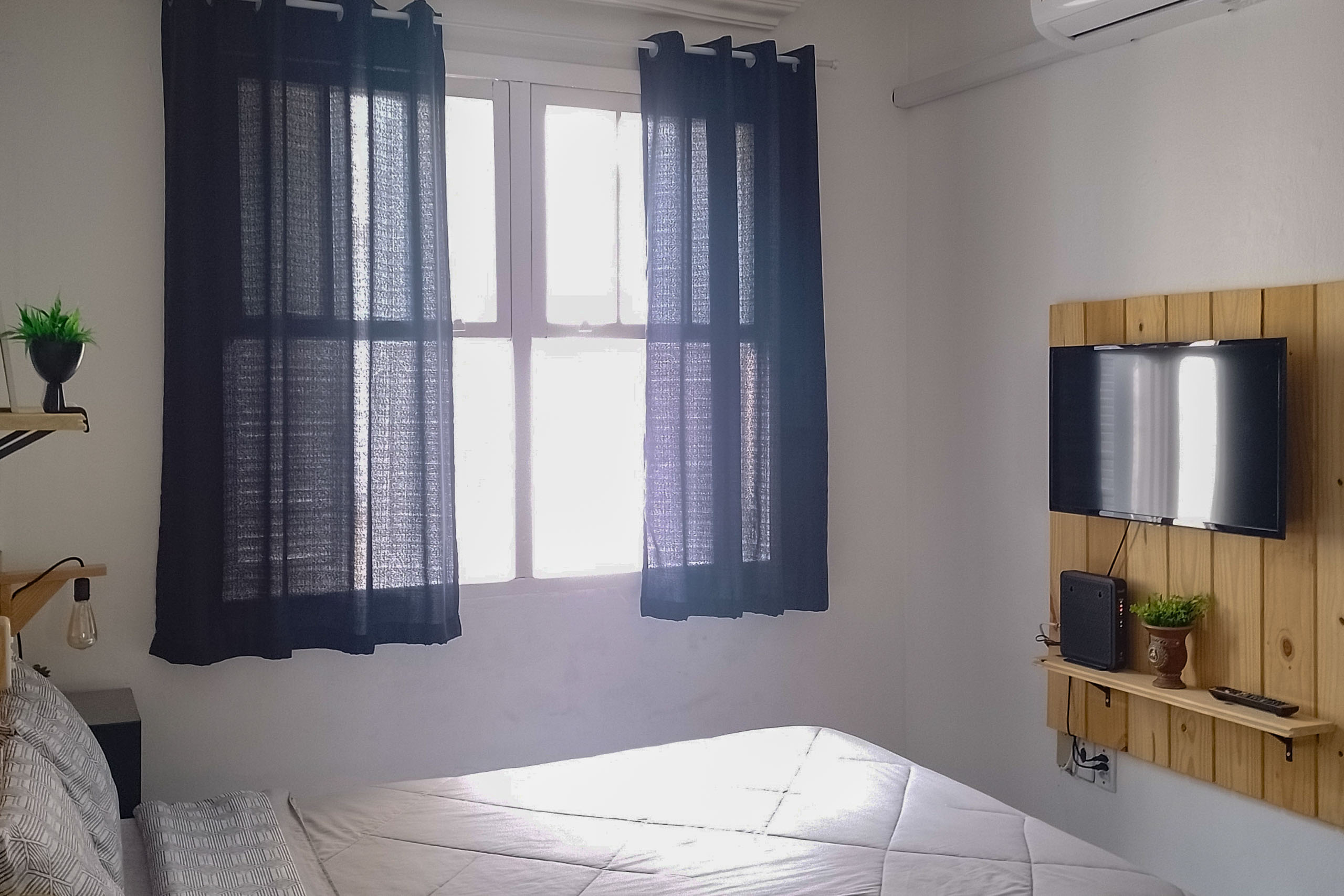 Bright light shines through dark blue curtains in a bedroom, with the white bed facing a small mounted TV. 