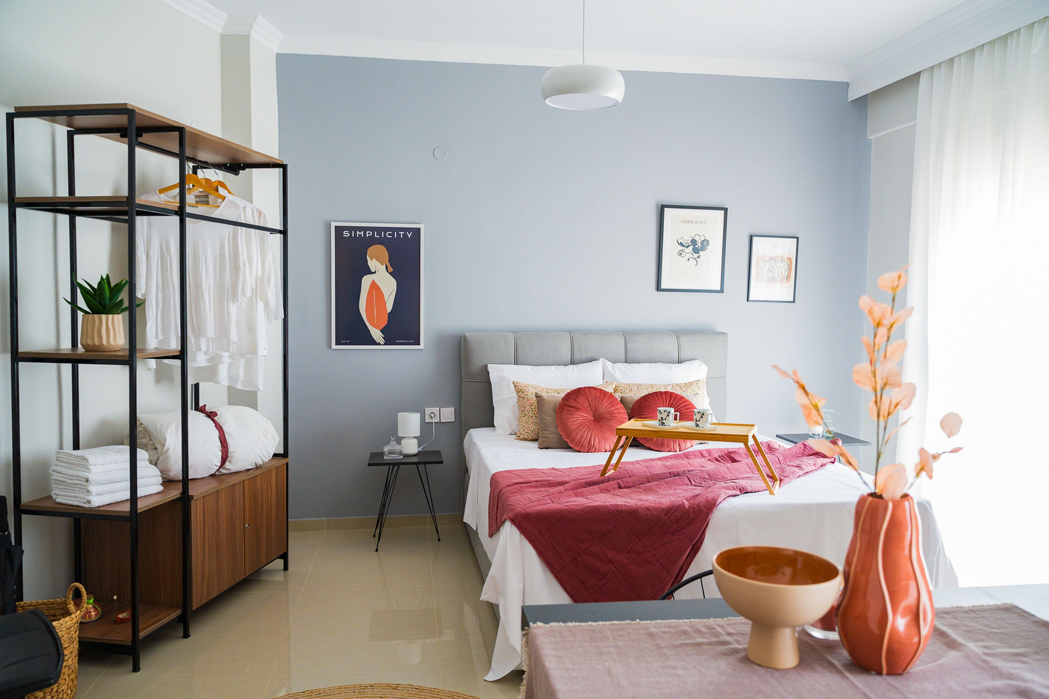 Modern bedroom with a sparse gallery wall, an exposed closet, artful decor and long white curtains letting in the sunlight. 