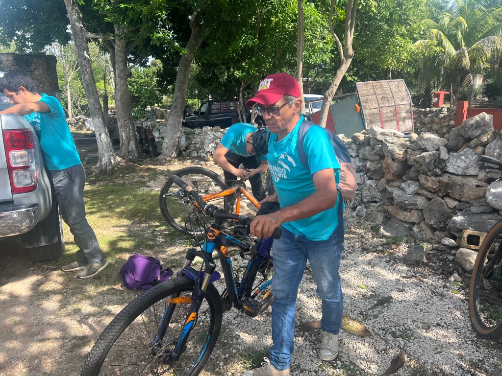 Man in a blue t-shirt and red baseball cap holds a bike while two other men prepare bikes in the background