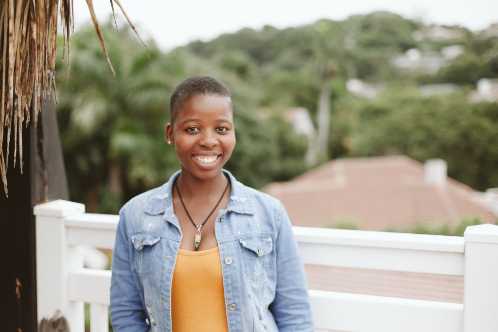 Woman with blue jean jacket and orange top stand in front a blurred backdrop