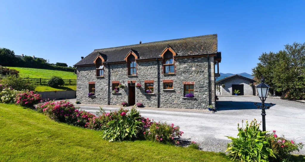 A picturesque stone house set against a backdrop of lush greenery and a clear blue sky. The house has two stories and is made of gray stone with a dark gray shingled roof.
A well-maintained garden with vibrant pink and red flowers borders the gravel driveway leading to the house.