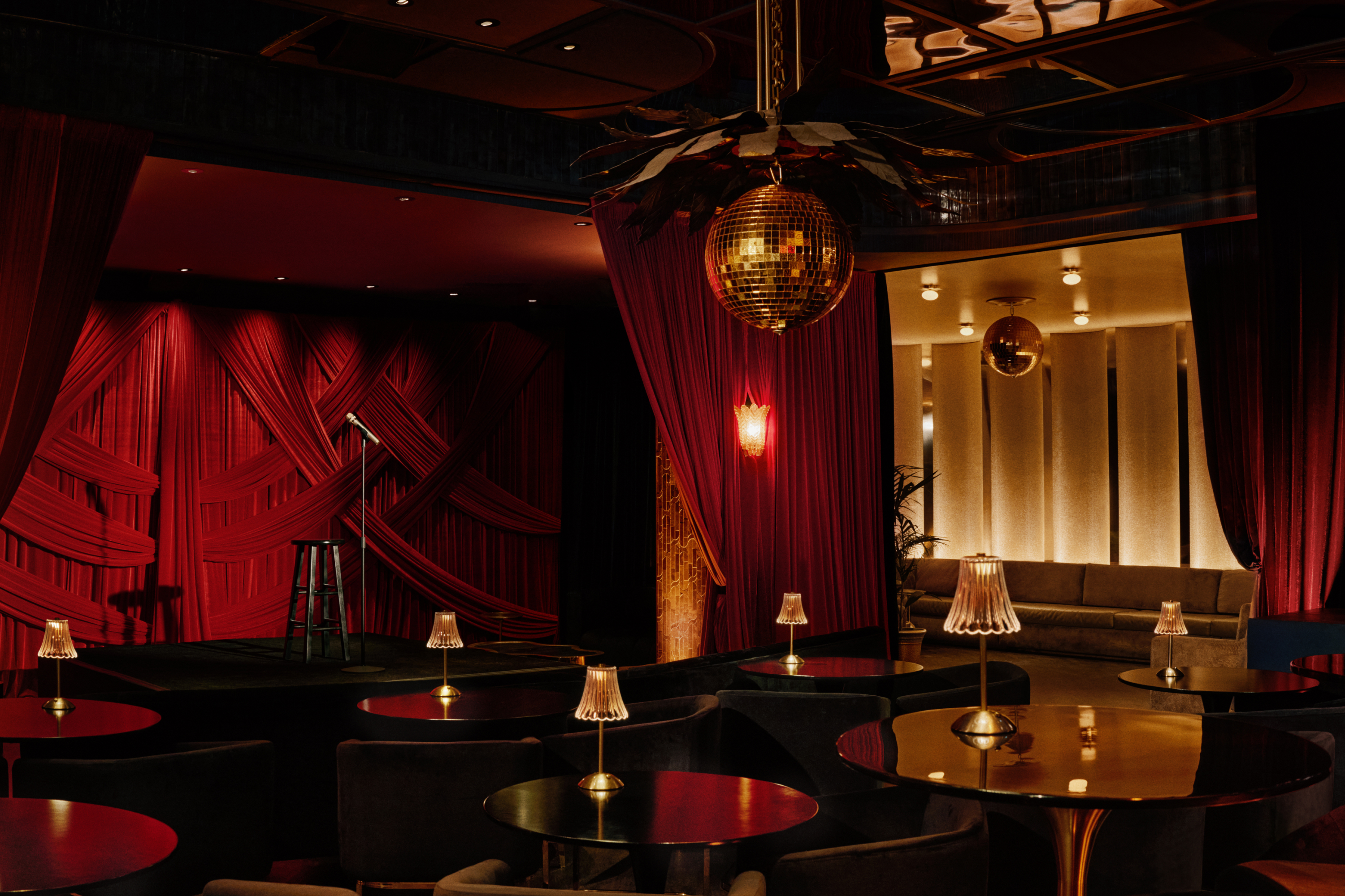 A dimly lit fancy room with plush red curtains, a stage, small round tables with small lamps and two arm chairs each, and a gold room with a gold couch in the back