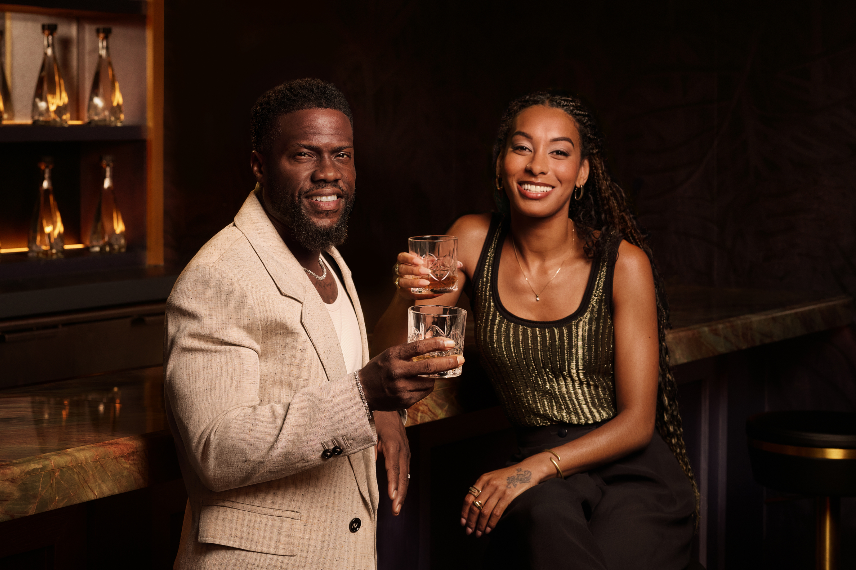 Kevin Hart and a woman smile at the camera while holding drinks at a bar