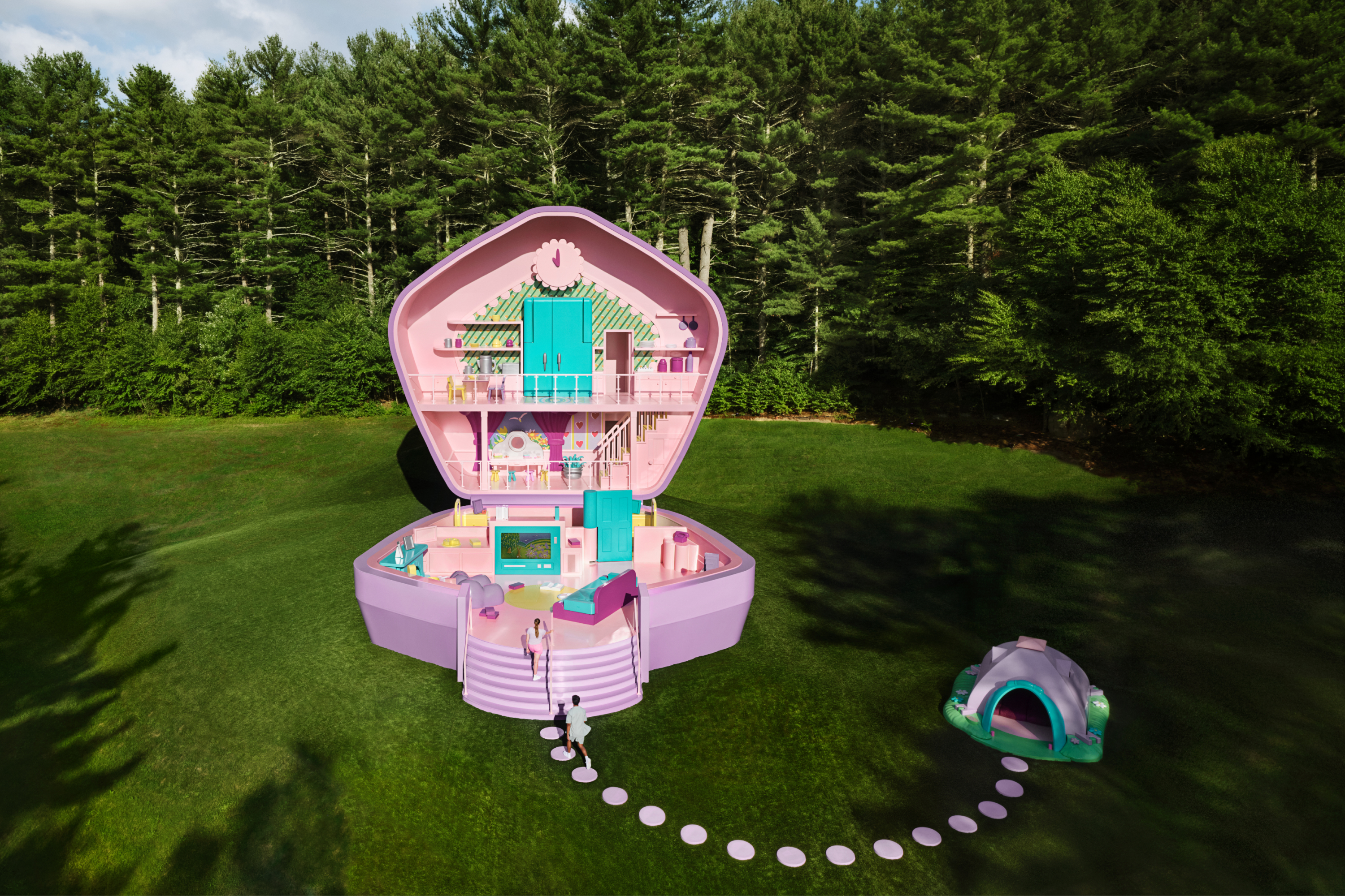 A life-sized purple Polly Pocket compact sits open in a lush green field with a forest in the background, with a life-sized Action Park Tent 10 feet away.