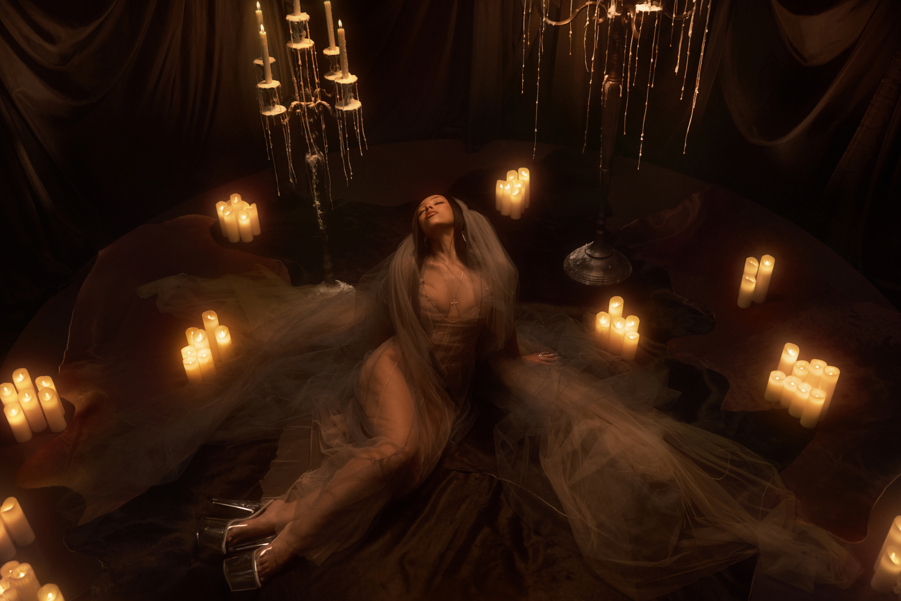 Doja Cat sits in the center of a small circular stage with a sheer lace veil and bodice, surrounded by dimly lit candles and candelabras with dripping wax.