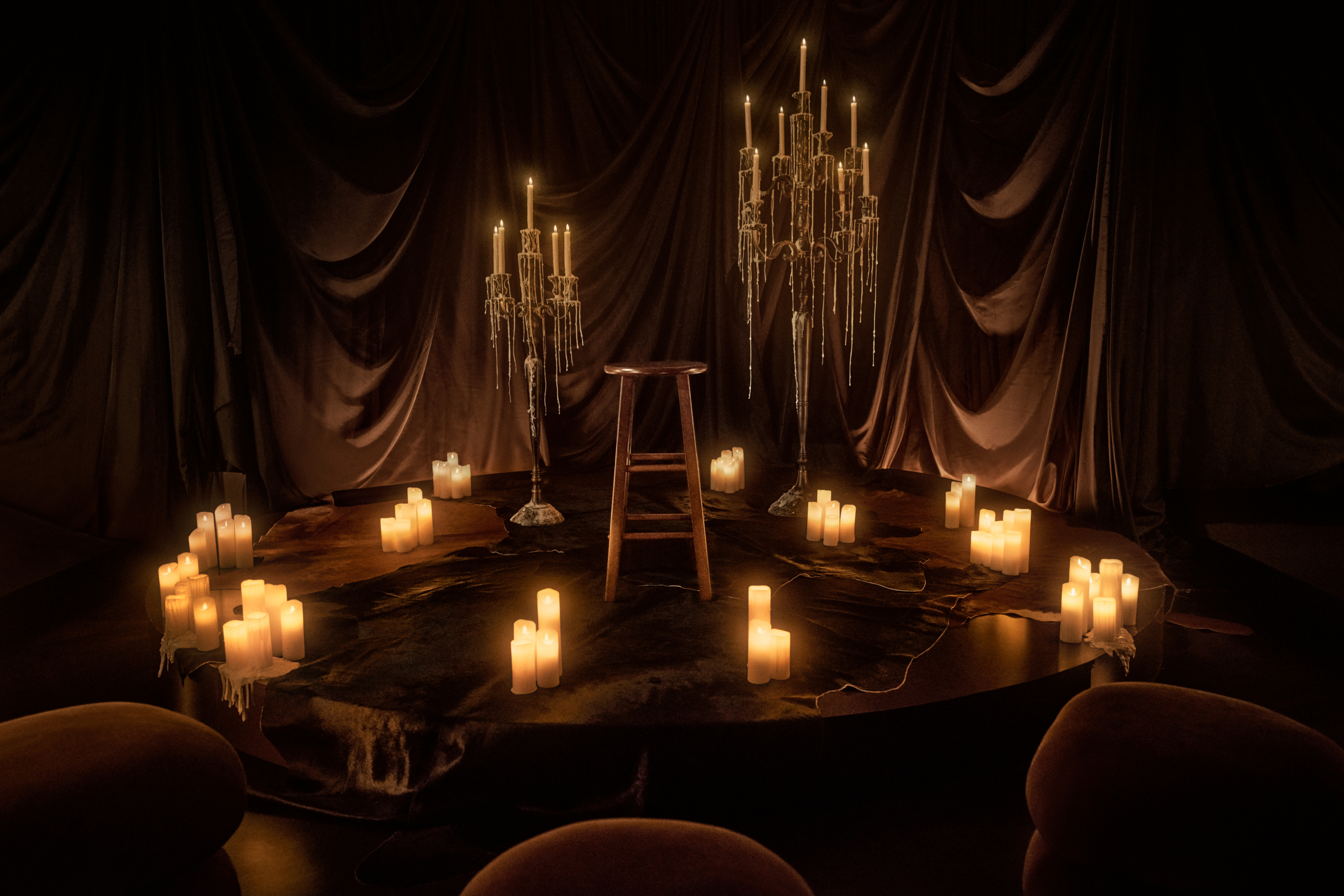 A small circular stage sits in the foreground with a single stool in the middle, surrounded by candles, cloaks and candelabras with a black curtain backdrop and audience seating in the foreground.