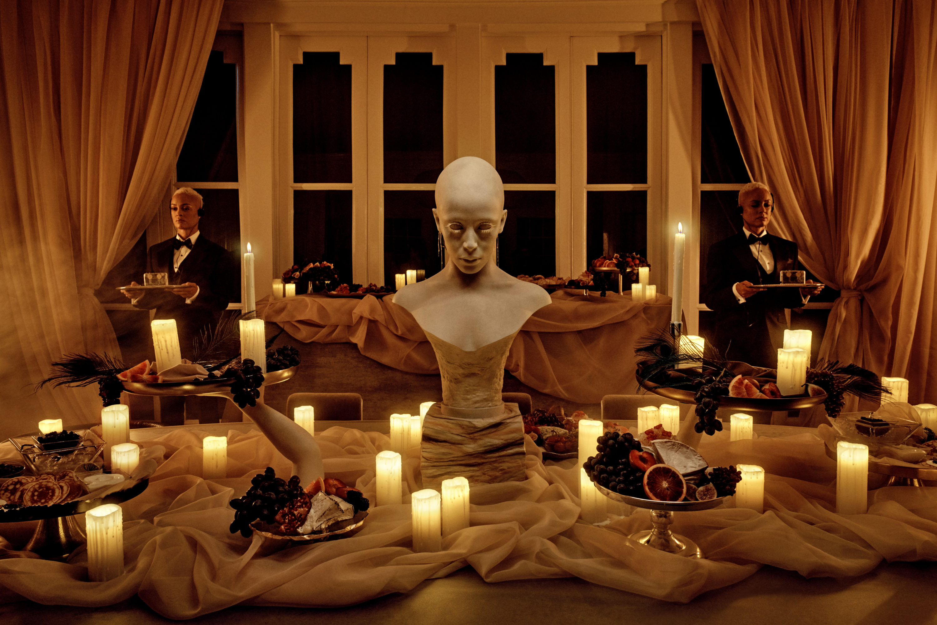 A marble bust resembling Doja Cat sits on a long table with gauze linens, candles and serving plates with fruts, caviar and cheese surrounding it. In the background, you can see a window framed by curtains and two servers in tuxedo uniforms holding serving platters.