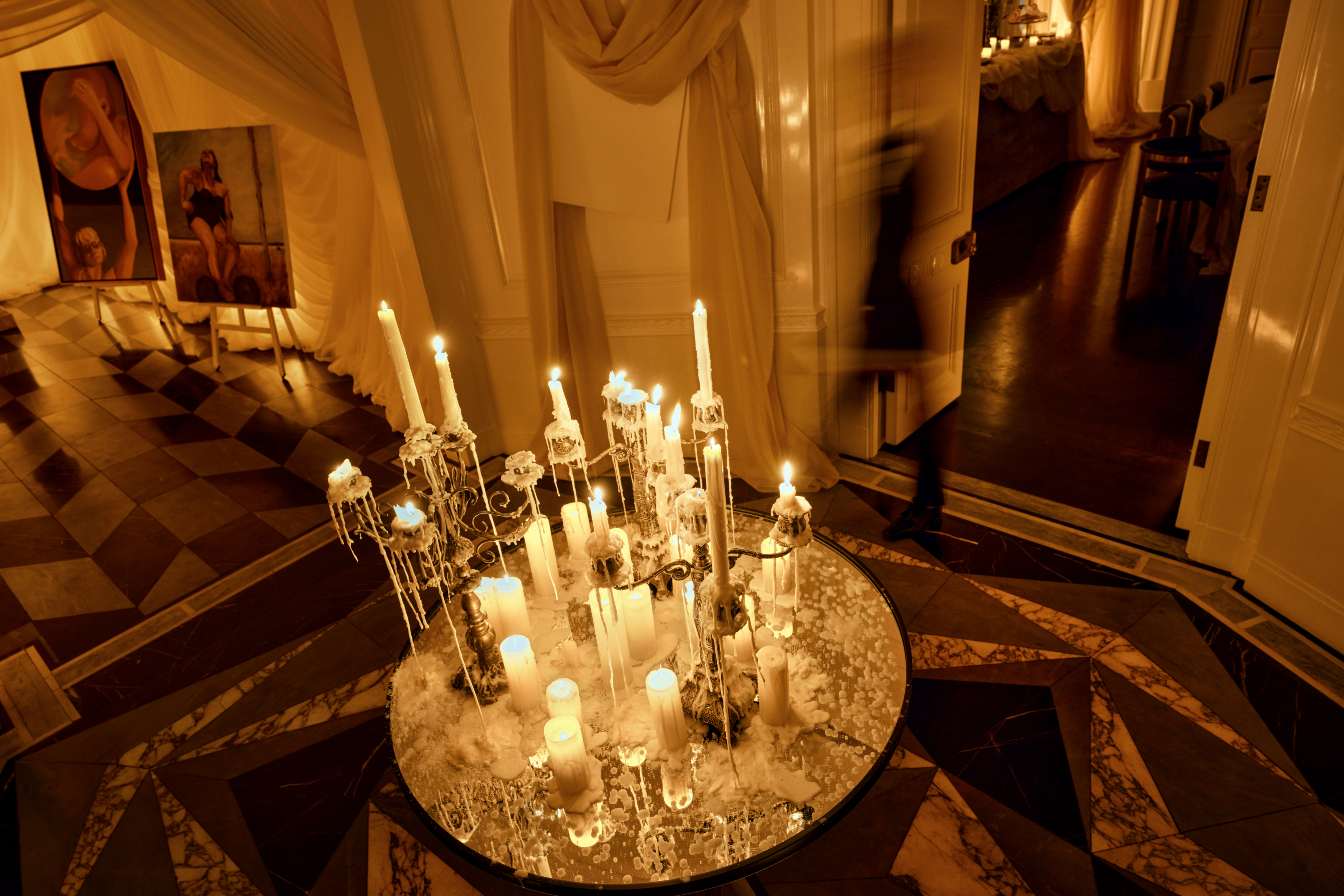 A series of candelabras with melting wax sit on a circular entry way table with entrances to two different rooms on either side. You can see a blurred shadow of a person walking through the room.