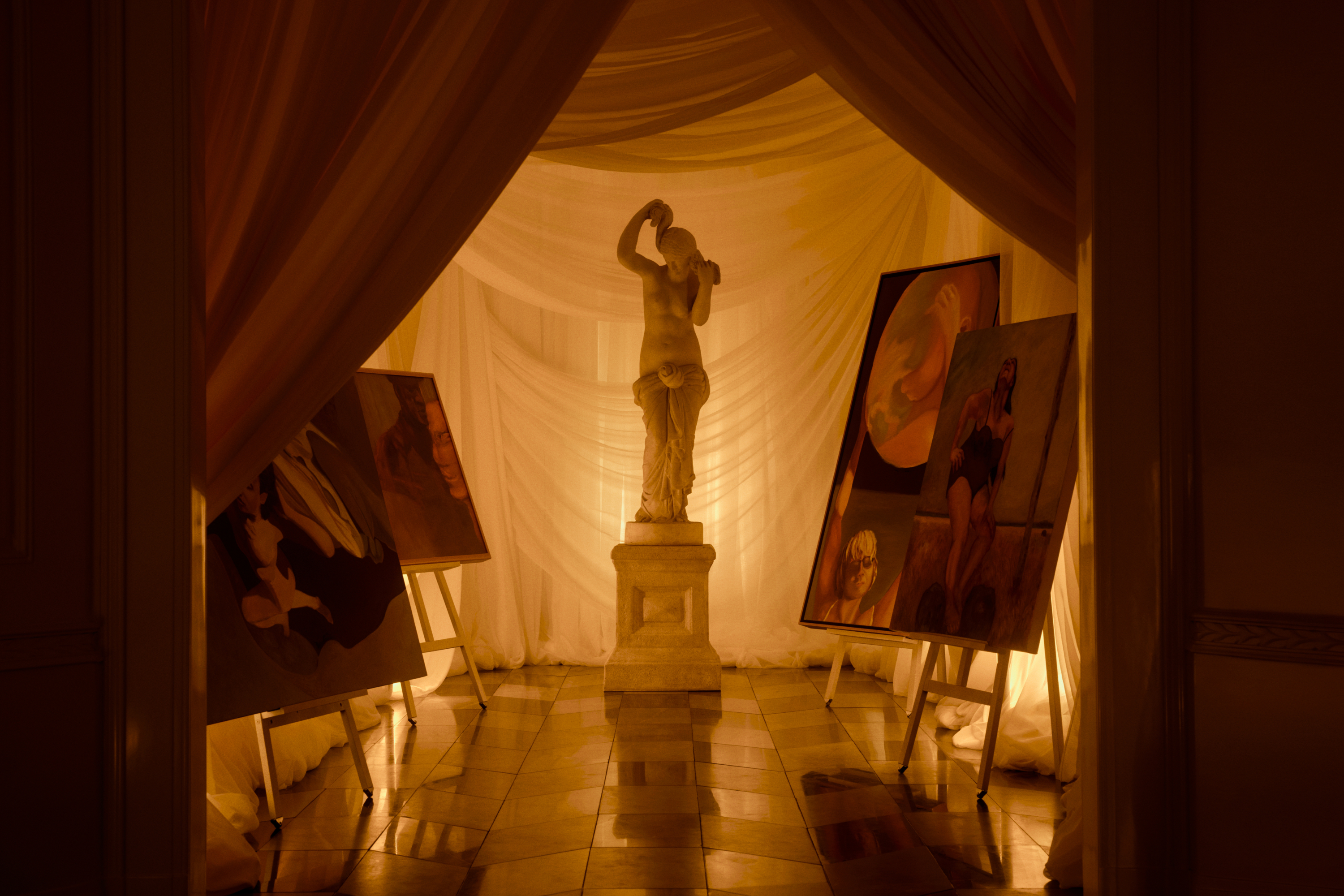 A marble sculpture of a person sits in the middle of a small gallery space with drapery all around and four different paintings resting on easels.