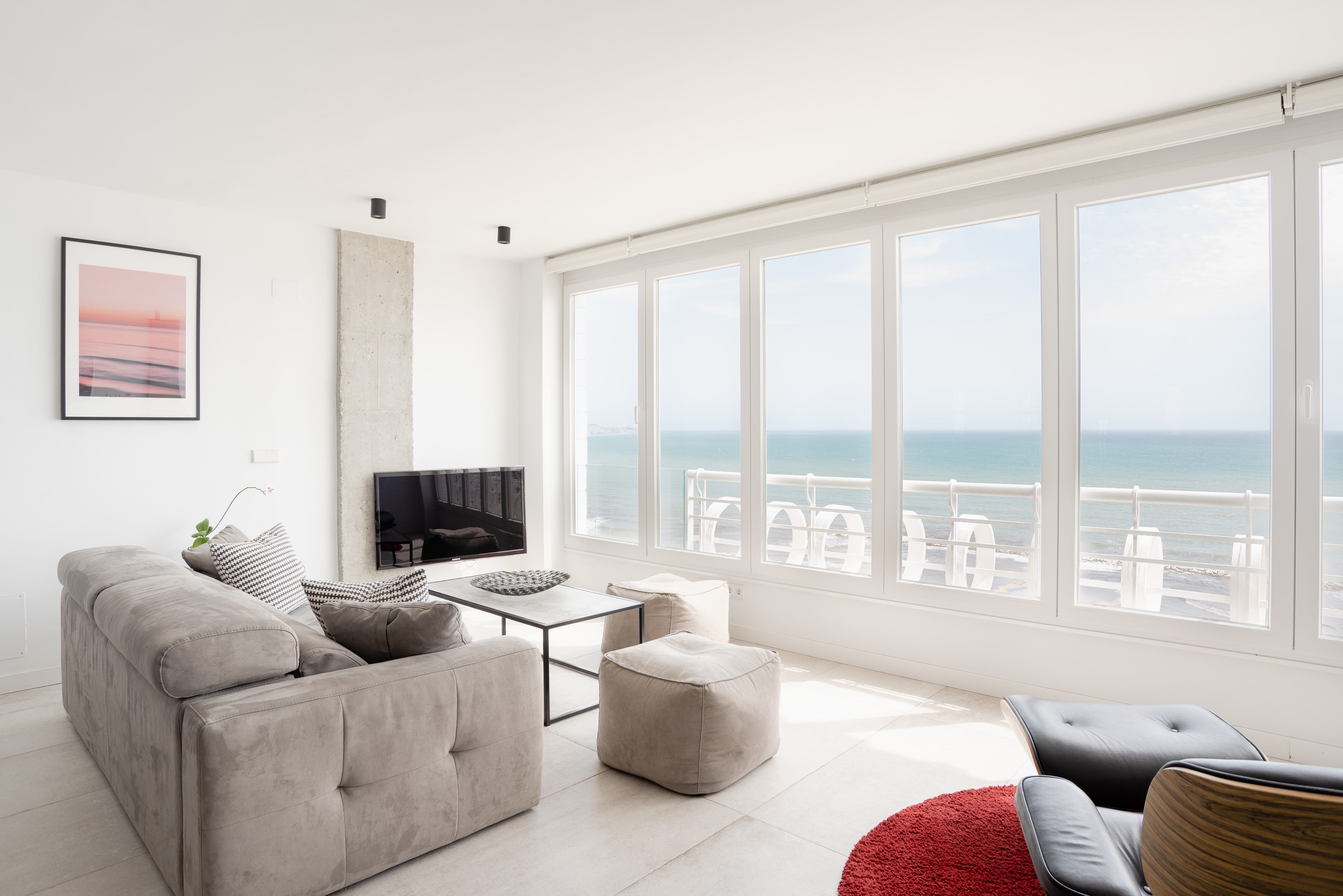A modern living room featuring a light gray sectional sofa with decorative pillows, a glass coffee table, and a flat-screen TV mounted on the wall. Large windows offer a stunning view of the ocean, with bright blue water and a clear sky. A small potted plant is on the table, and a red circular rug adds a pop of color to the floor. A concrete pillar is visible between the sofa and the windows, enhancing the contemporary aesthetic of the space.