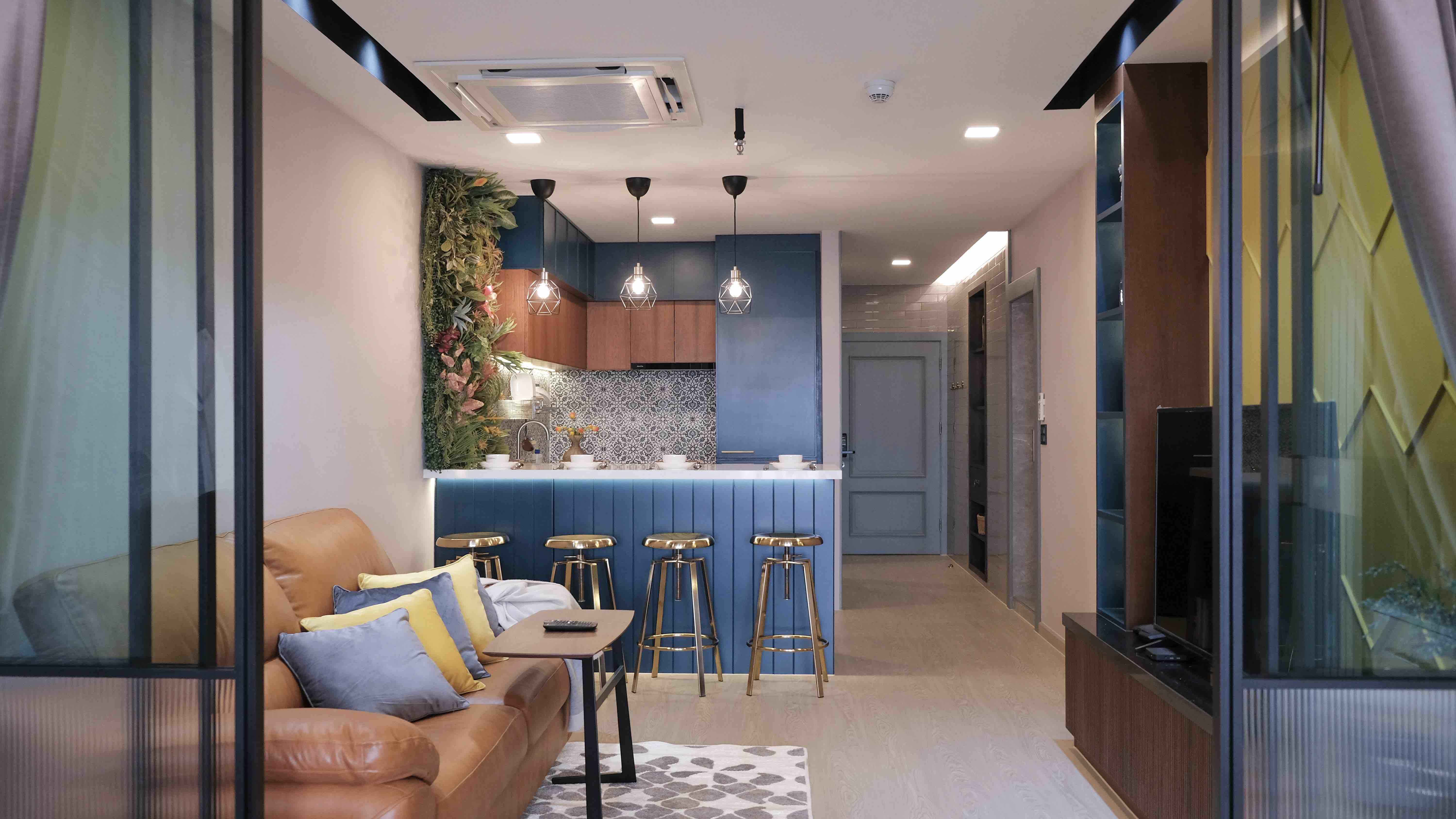 A modern living space featuring a stylish brown leather sofa adorned with colorful cushions in shades of blue and yellow. The kitchen area, visible in the background, boasts blue cabinetry, geometric pendant lights, and a patterned backsplash. A small dining table with golden bar stools is positioned at the kitchen counter, and a glass partition separates the living area from the hallway. The room is well-lit with natural light streaming in, creating a warm and inviting atmosphere.