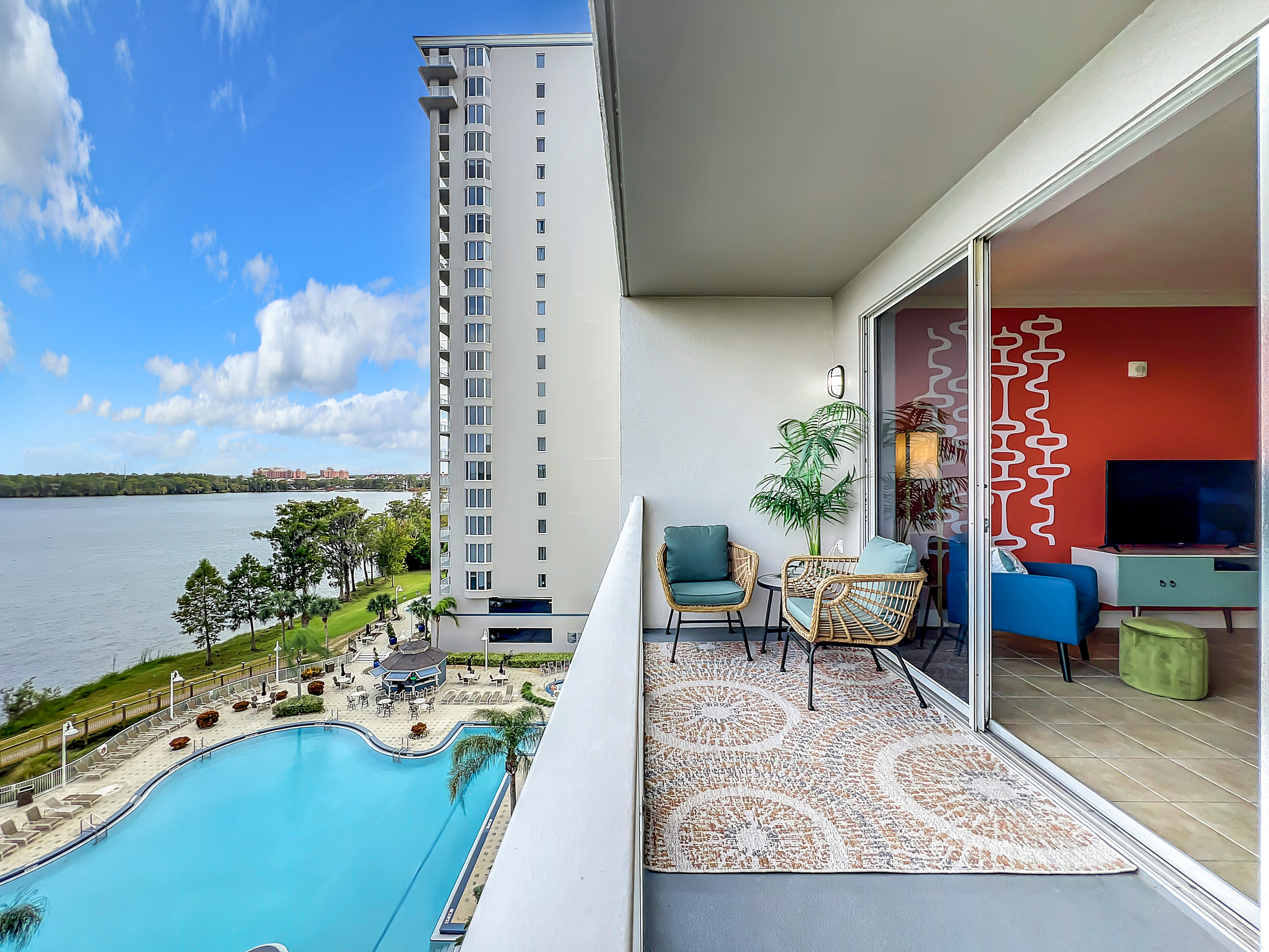 A balcony view overlooking a pool and body of water. The balcony features two woven chairs and a small table. Inside, a cozy living room with a blue couch, a green ottoman, and a television can be seen through sliding glass doors. The sky is partly cloudy.