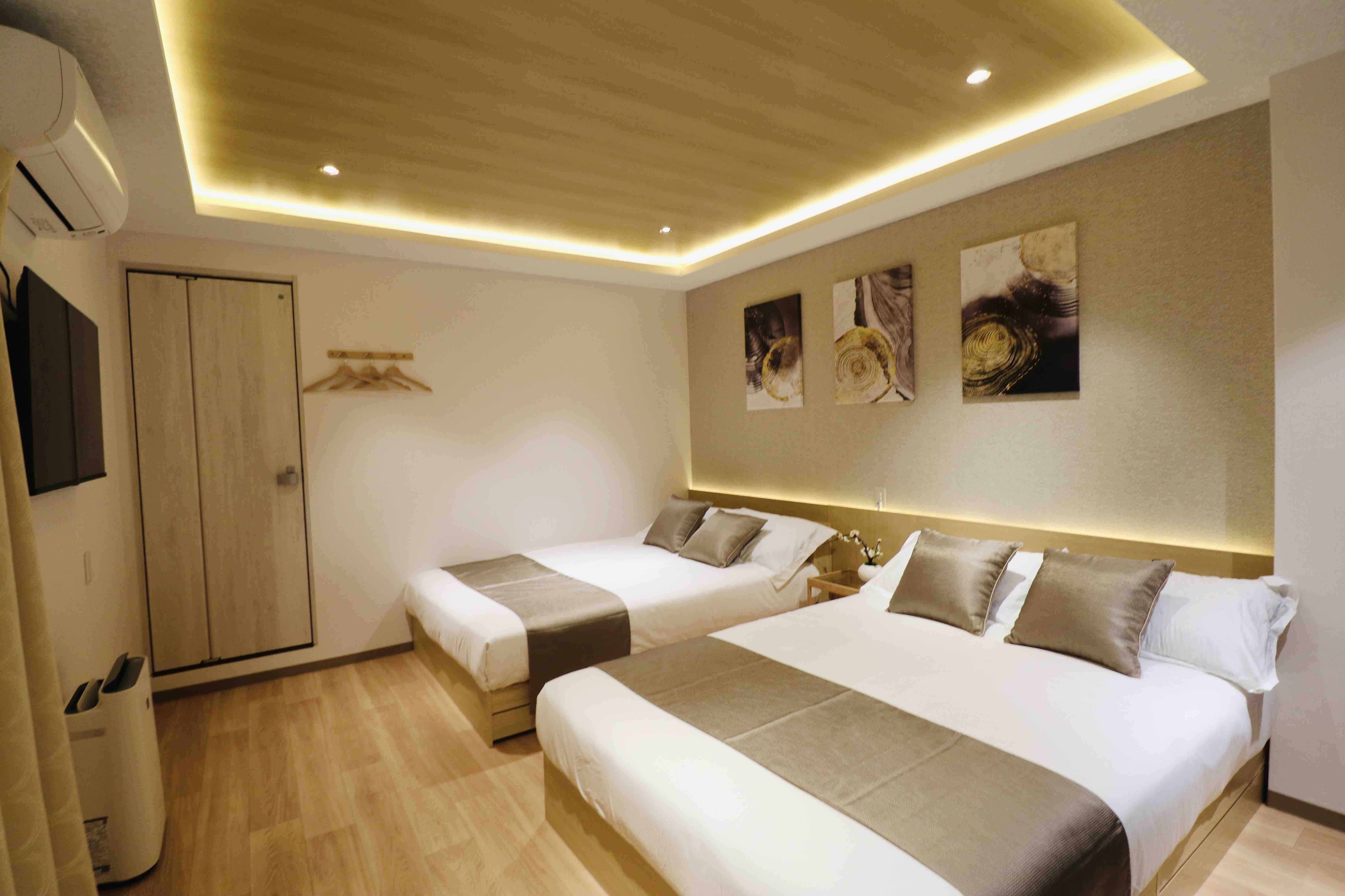 A modern bedroom featuring two neatly made beds with gray and white bedding. The room has a warm ambiance with recessed lighting along the ceiling. On the wall, there are three abstract art pieces. A flat-screen TV is mounted on the wall opposite the beds, and a wooden closet door is visible. A coat rack with wooden hangers is mounted beside the closet, and there is a small white drawer beside one of the beds.