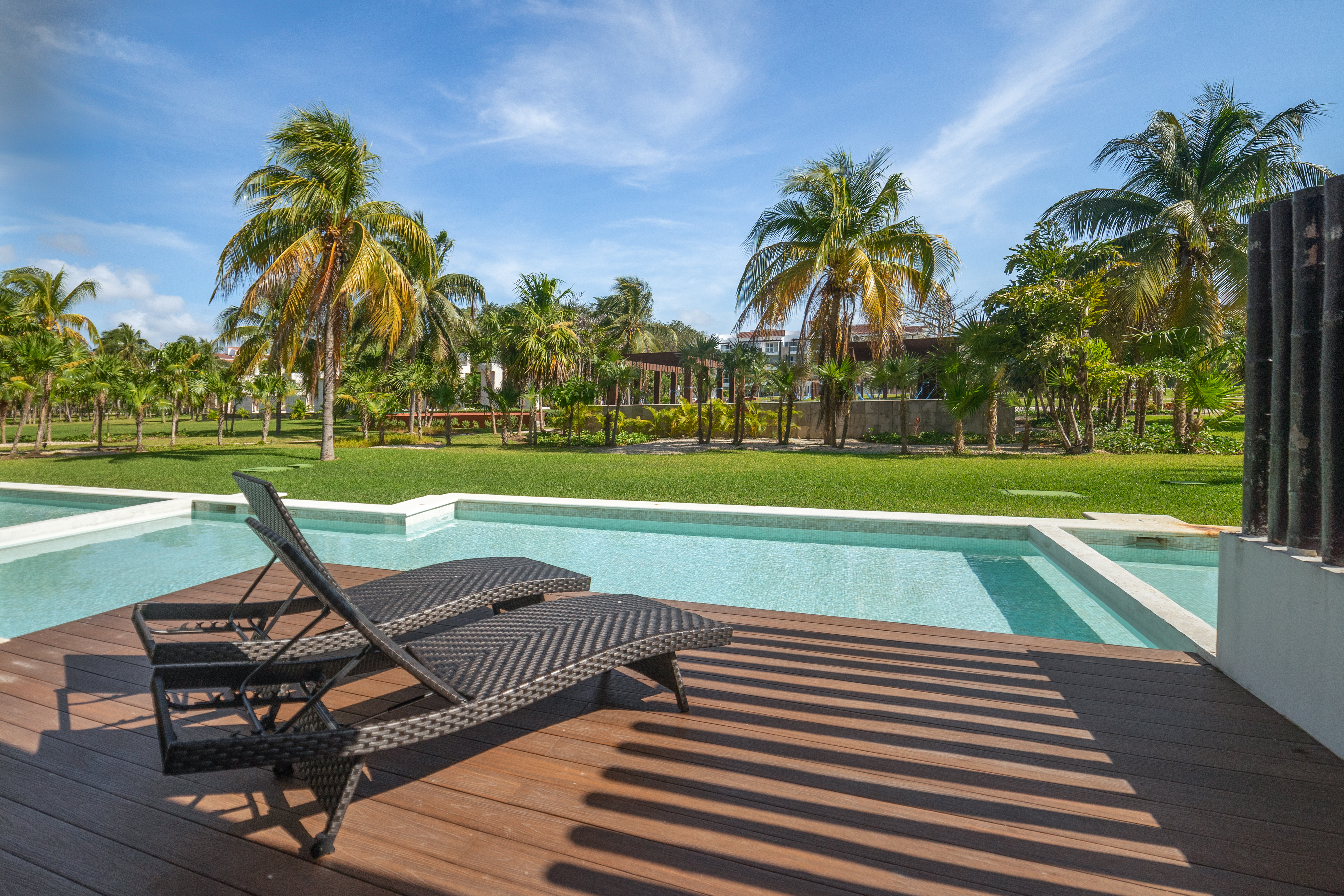 A serene outdoor patio featuring a swimming pool surrounded by lush greenery and palm trees. In the foreground, two black wicker lounge chairs are positioned on a wooden deck, facing the pool and the vibrant landscape beyond under a clear blue sky.