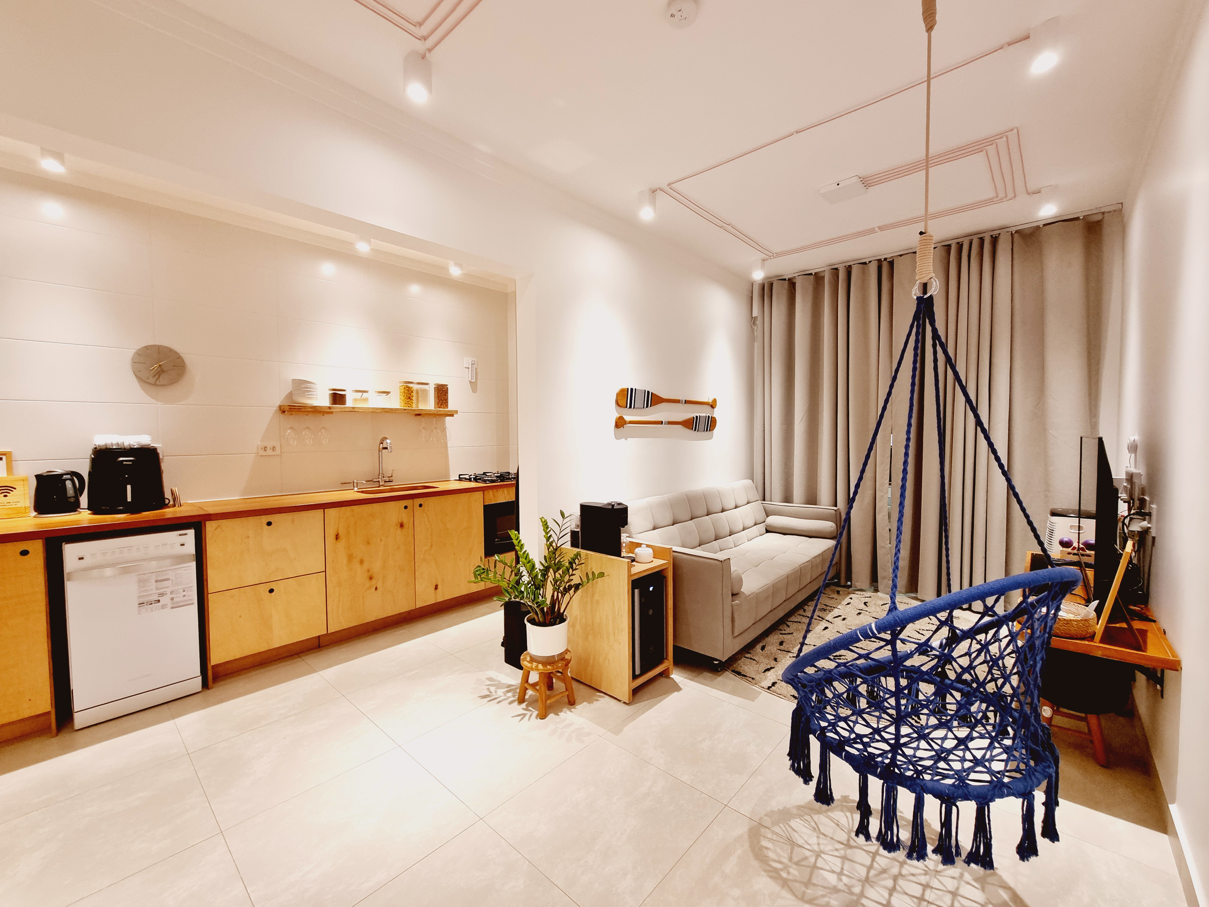 A cozy and modern living space featuring a compact kitchen with wooden cabinets and a countertop, equipped with a black toaster, a coffee maker, a sink, and a gas stove. On one side, there's a gray sofa facing a small dining area with a wooden chair and a television. A unique blue macrame swing chair hangs from the ceiling, adding a playful touch to the room. The walls are adorned with light-colored curtains, and decorative elements like a clock and decorative oars on the wall enhance the aesthetic.