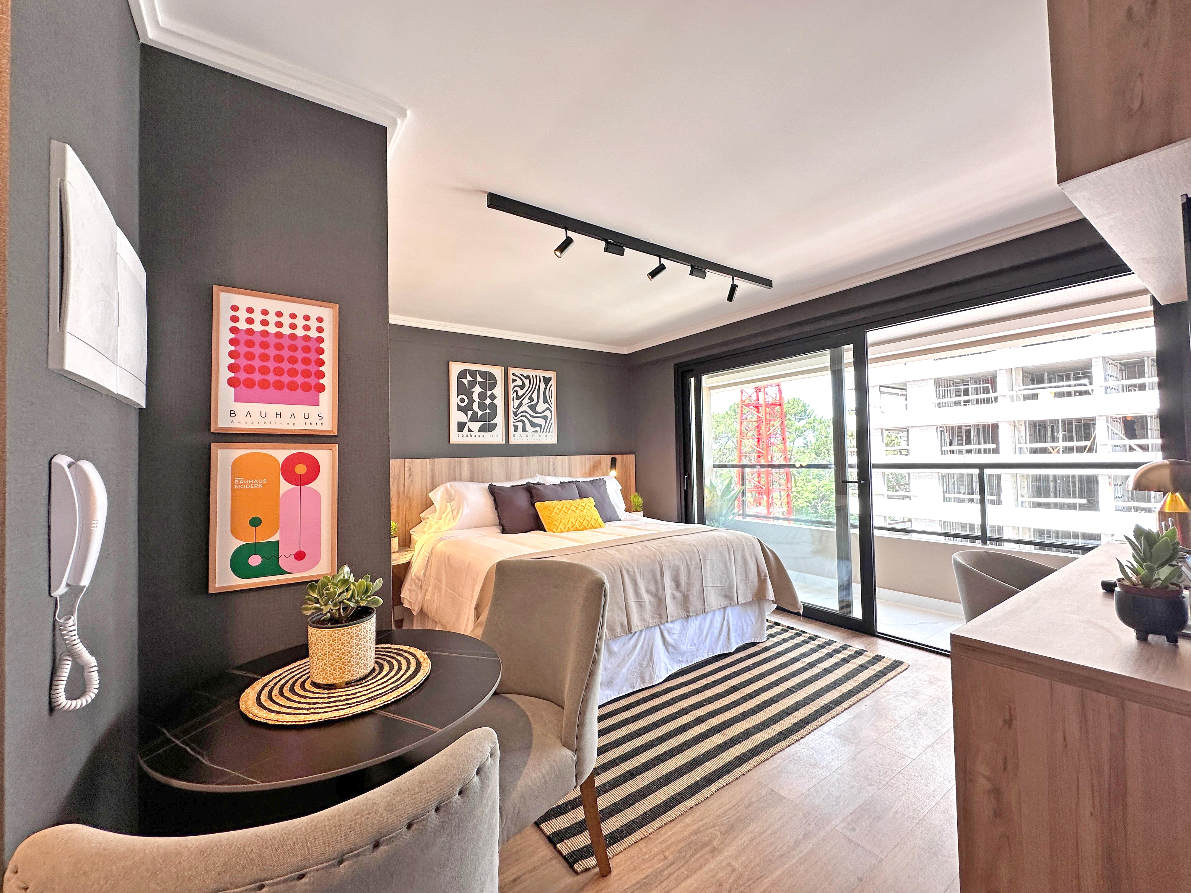 A modern bedroom featuring a large bed with decorative pillows and a light-colored blanket. The walls are painted dark gray, adorned with colorful art prints. A small round black table with a woven placemat and a potted plant sits beside a plush chair. To the right, a wooden dresser holds another small plant. The room has large glass doors leading to a balcony.