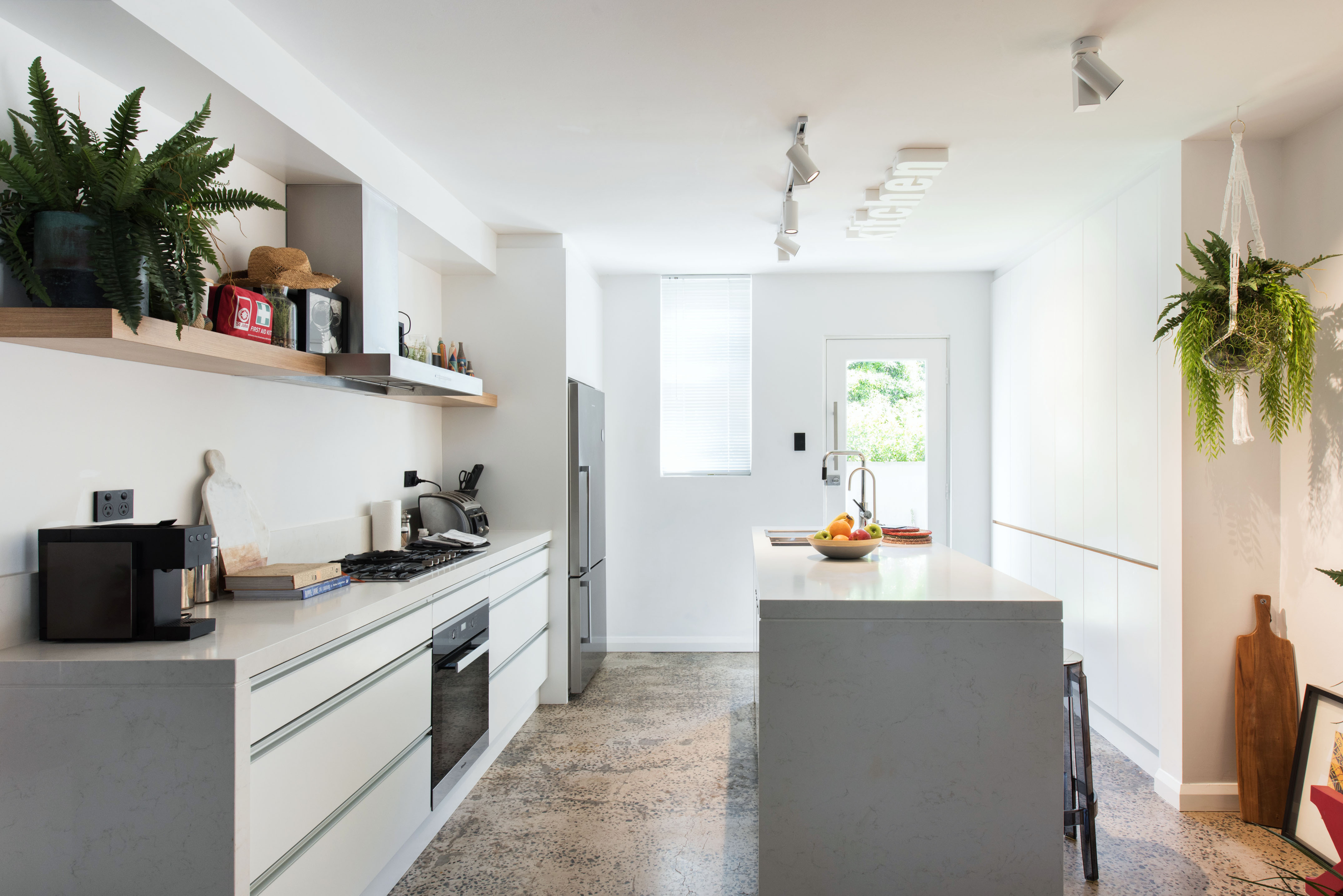 A modern kitchen featuring a clean, minimalist design. The kitchen has white cabinets, a large island with a marble countertop, and a stainless steel stove. Above the stove, there is a sleek hood, and a shelf displays various decorative items and kitchen essentials. A bowl of colorful fruits sits on the island. The space is well-lit with track lighting and natural light coming from a window, and there are hanging plants adding a touch of greenery. A door leads outside.