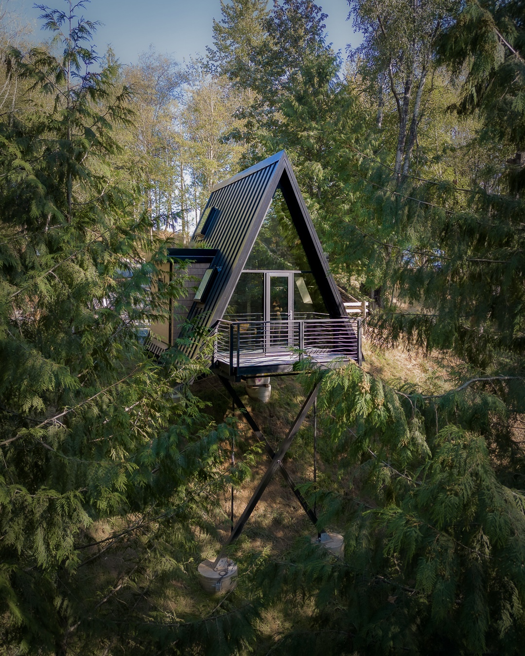 A modern A-frame house perched among trees, featuring a black exterior with large glass windows. The house is elevated on stilts, with a balcony visible, surrounded by lush greenery and a clear blue sky above.