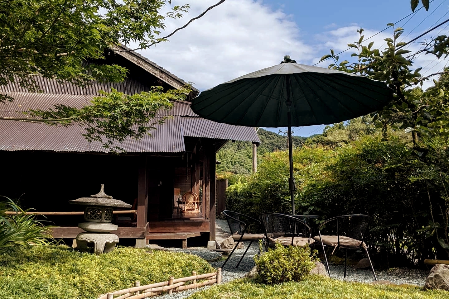 A serene outdoor scene featuring a traditional wooden house with a corrugated roof, surrounded by lush greenery. In the foreground, there is a stone lantern and a small garden area with a green umbrella shading a couple of black metal chairs. The backdrop showcases a clear blue sky with a few clouds, adding to the peaceful atmosphere.